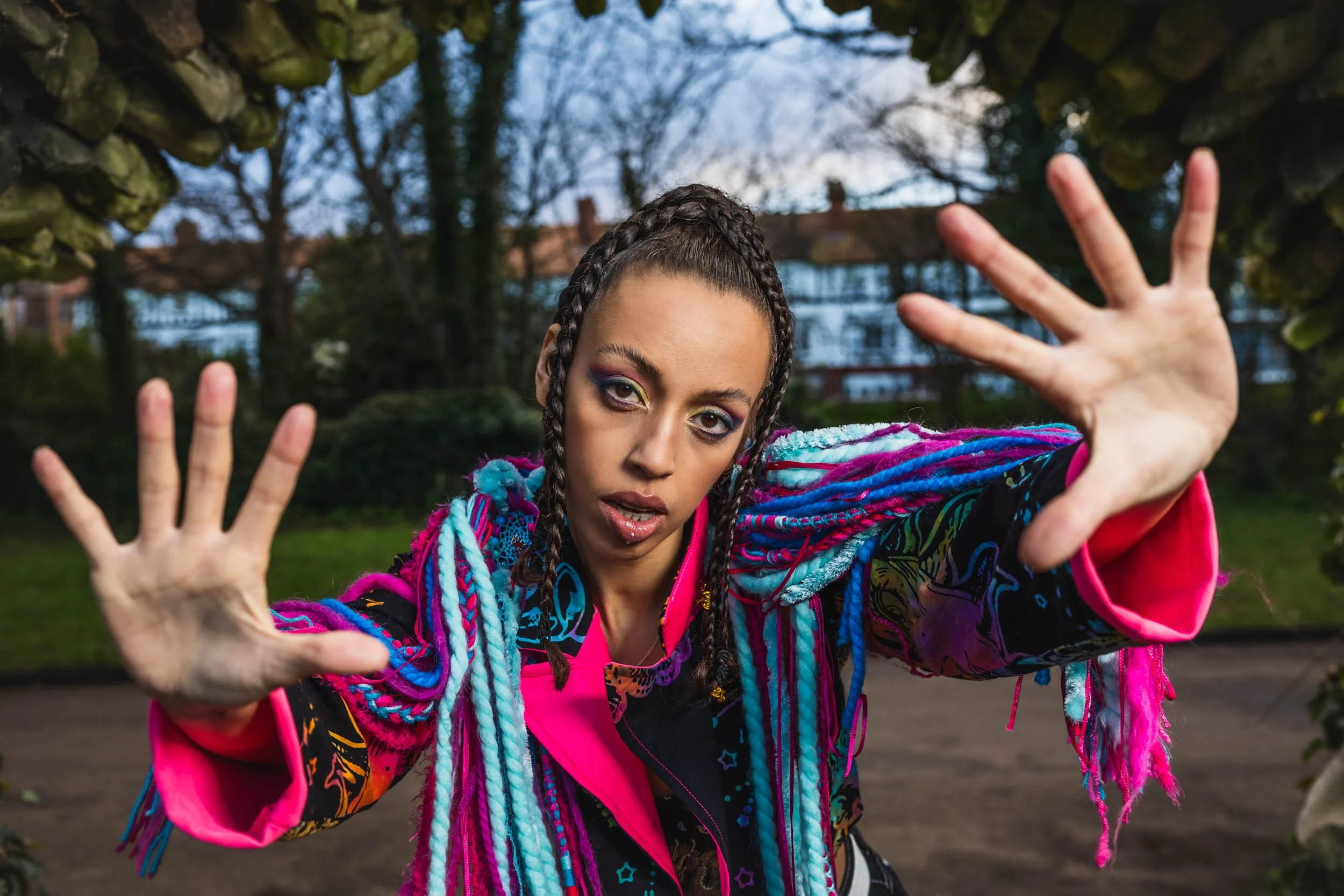 A woman with braided hair wearing a colorful jacket reaching out her hands toward the camera outdoors during daytime.