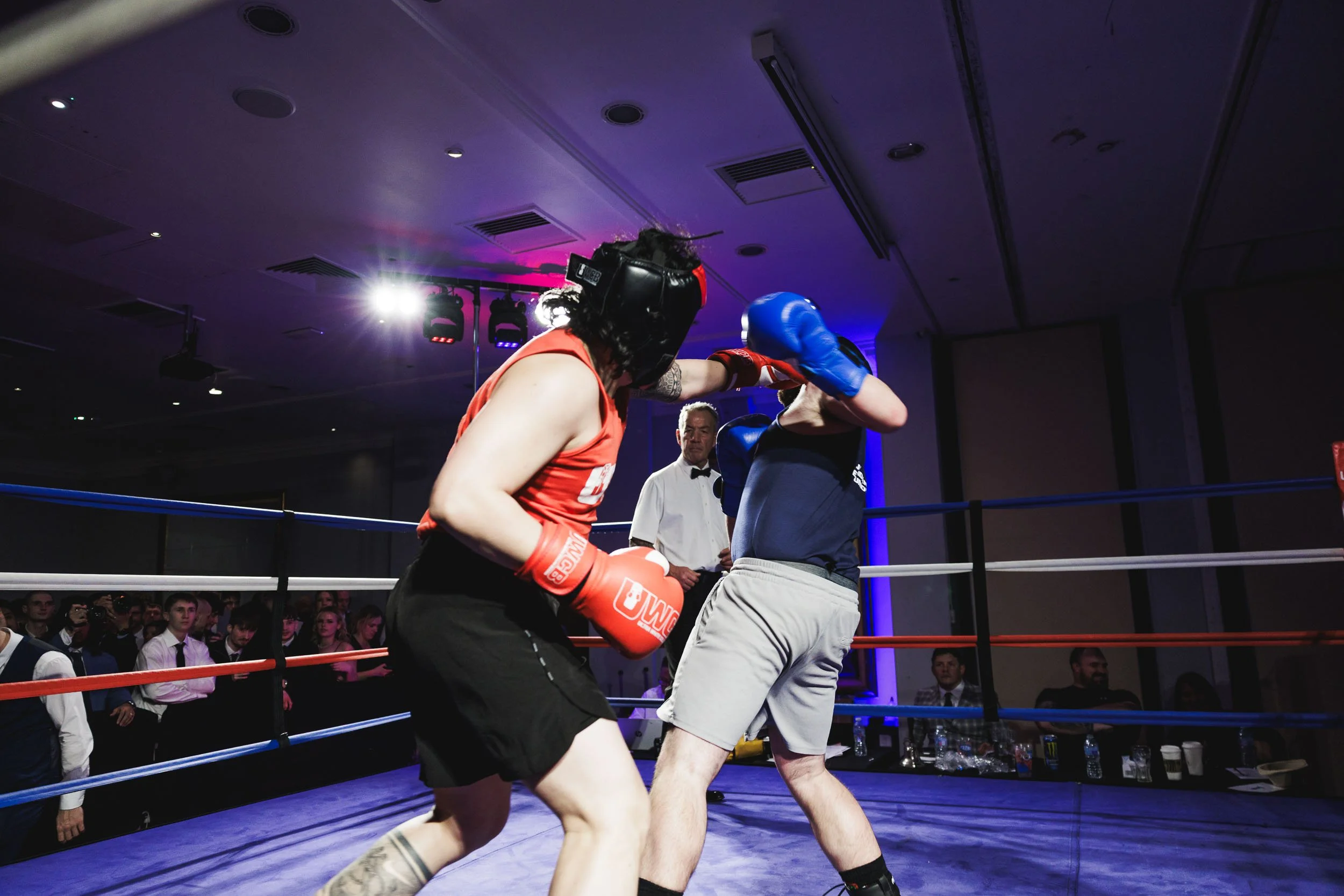 Two women boxing in a ring with an audience watching. One woman is wearing a black helmet, red gloves, red tank top, and black shorts. The other woman is wearing a blue helmet, blue gloves, dark shirt, and gray shorts. A referee is observing in the b