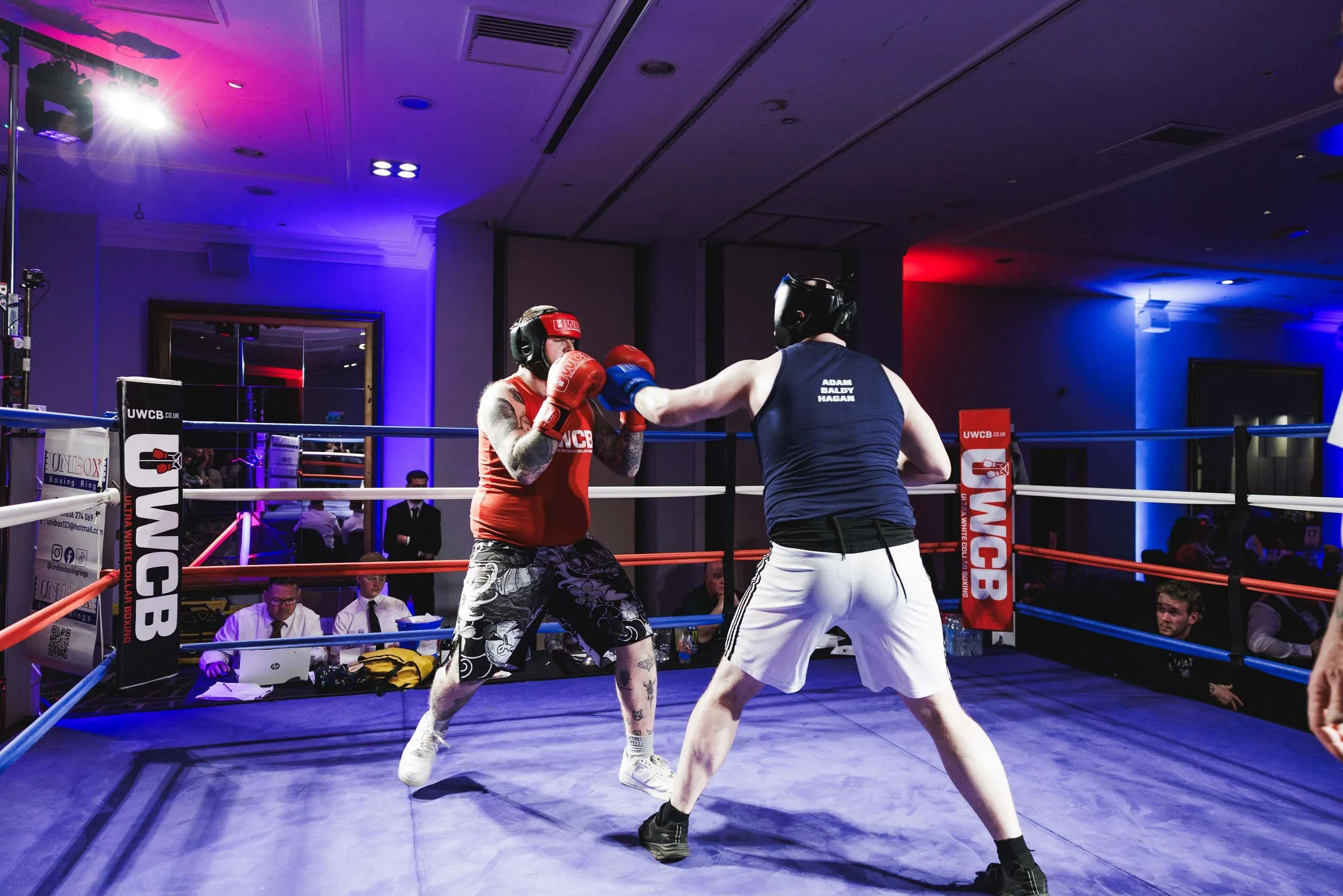 Boxing match between two male fighters in a ring, with one throwing a punch at the other, in a brightly lit indoor arena.