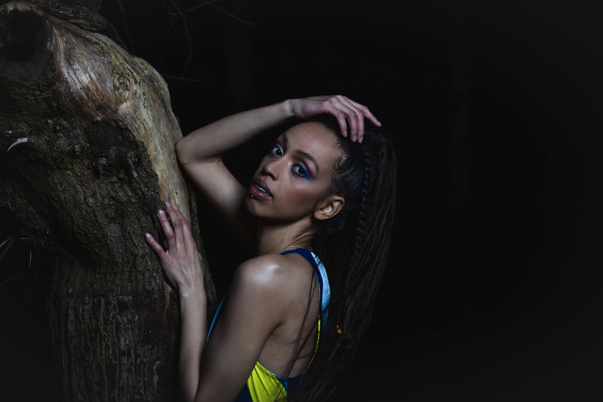 A woman with braided hair and colorful makeup posing beside a tree trunk in a dark environment