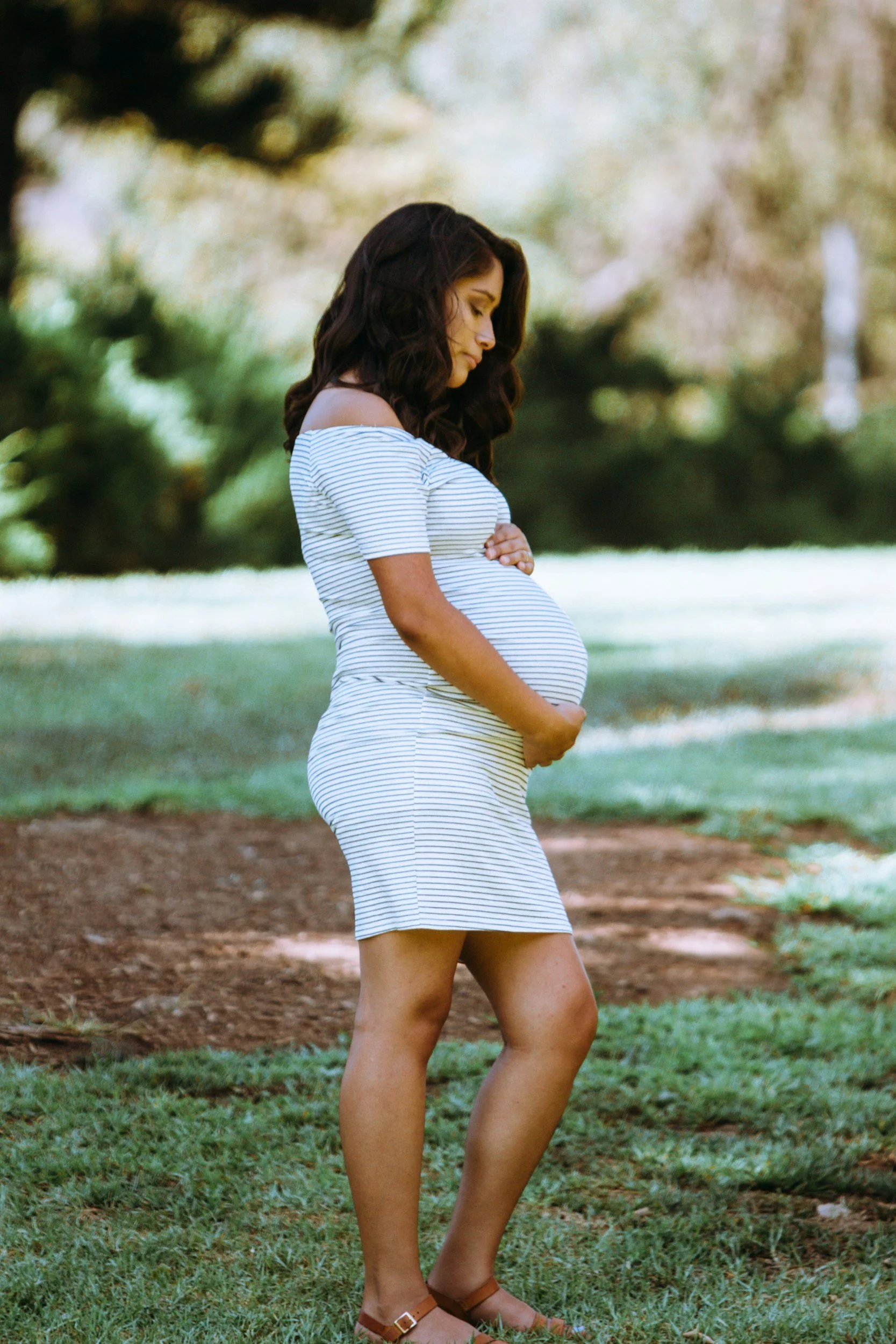 A pregnant woman in a striped dress standing outdoors on a grassy area, gently holding her belly, with trees in the background.