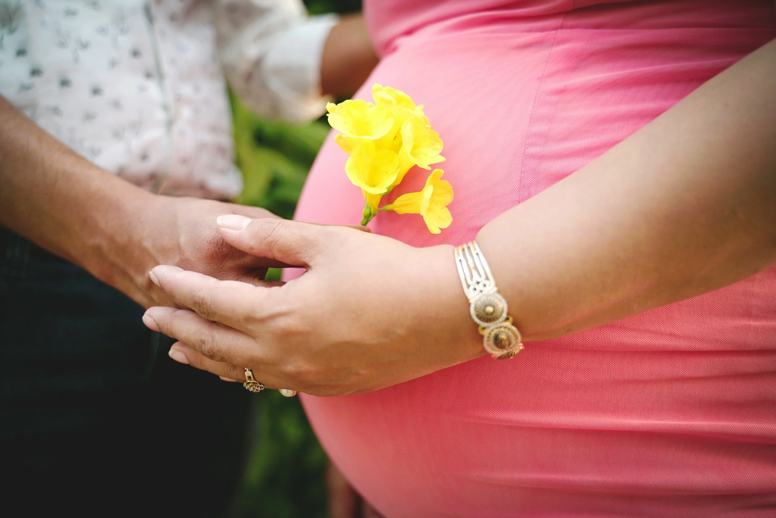 A pregnant woman in a pink dress holding a yellow flower, with her hand touching another person's hand.