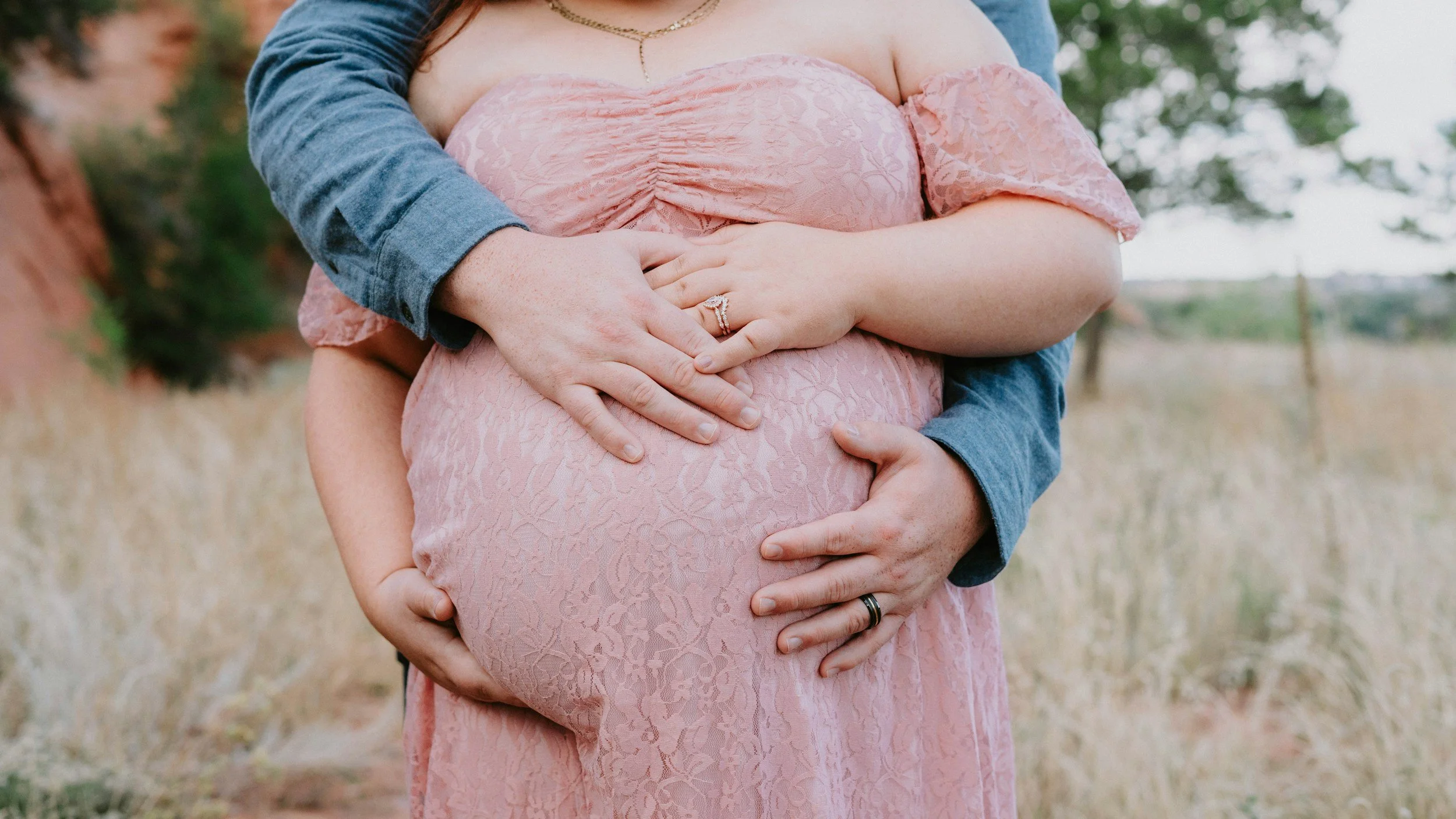Close-up of a pregnant woman wearing a pink lace dress with her partner’s hands gently holding her belly from above and below, set outdoors with field and trees.