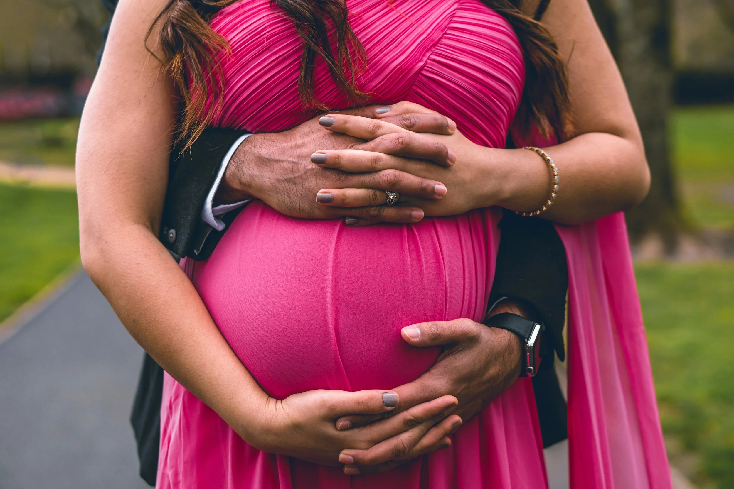 Close-up of a pregnant woman in a pink dress, with her partner's and partner's hand visible on her belly, outdoors in a park setting.