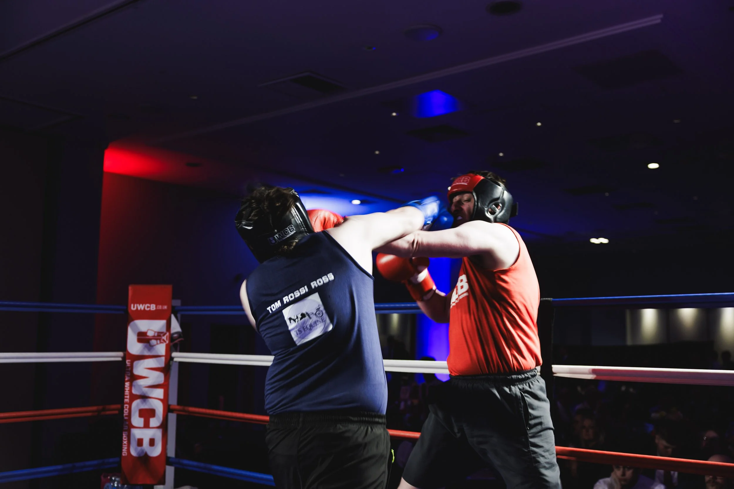 Two male boxers wearing headgear and boxing gloves fight in a ring, with one delivering a punch to the other's face.