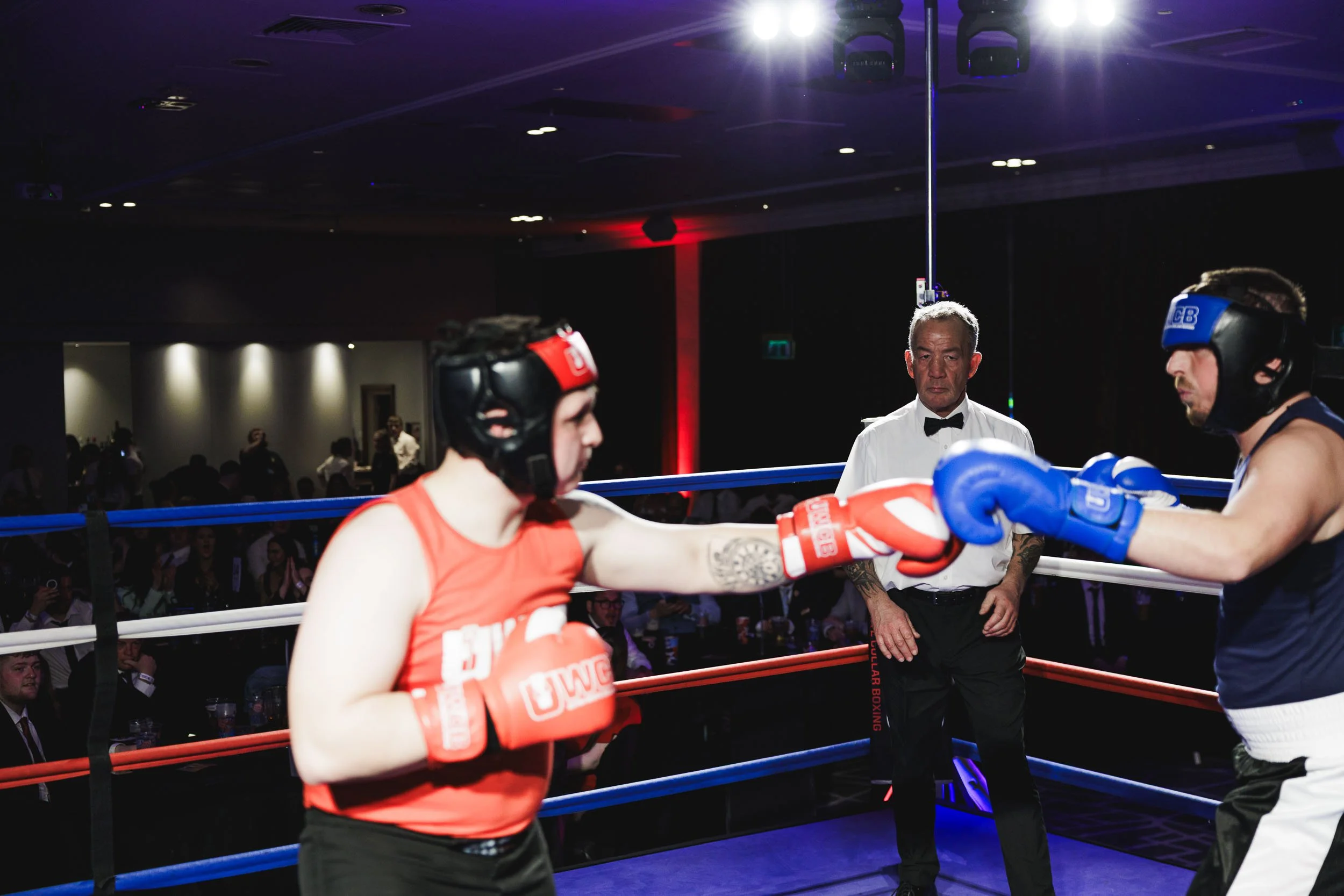 Women boxing match in a ring with referee observing, two fighters wearing protective headgear and gloves, one in red, the other in blue.