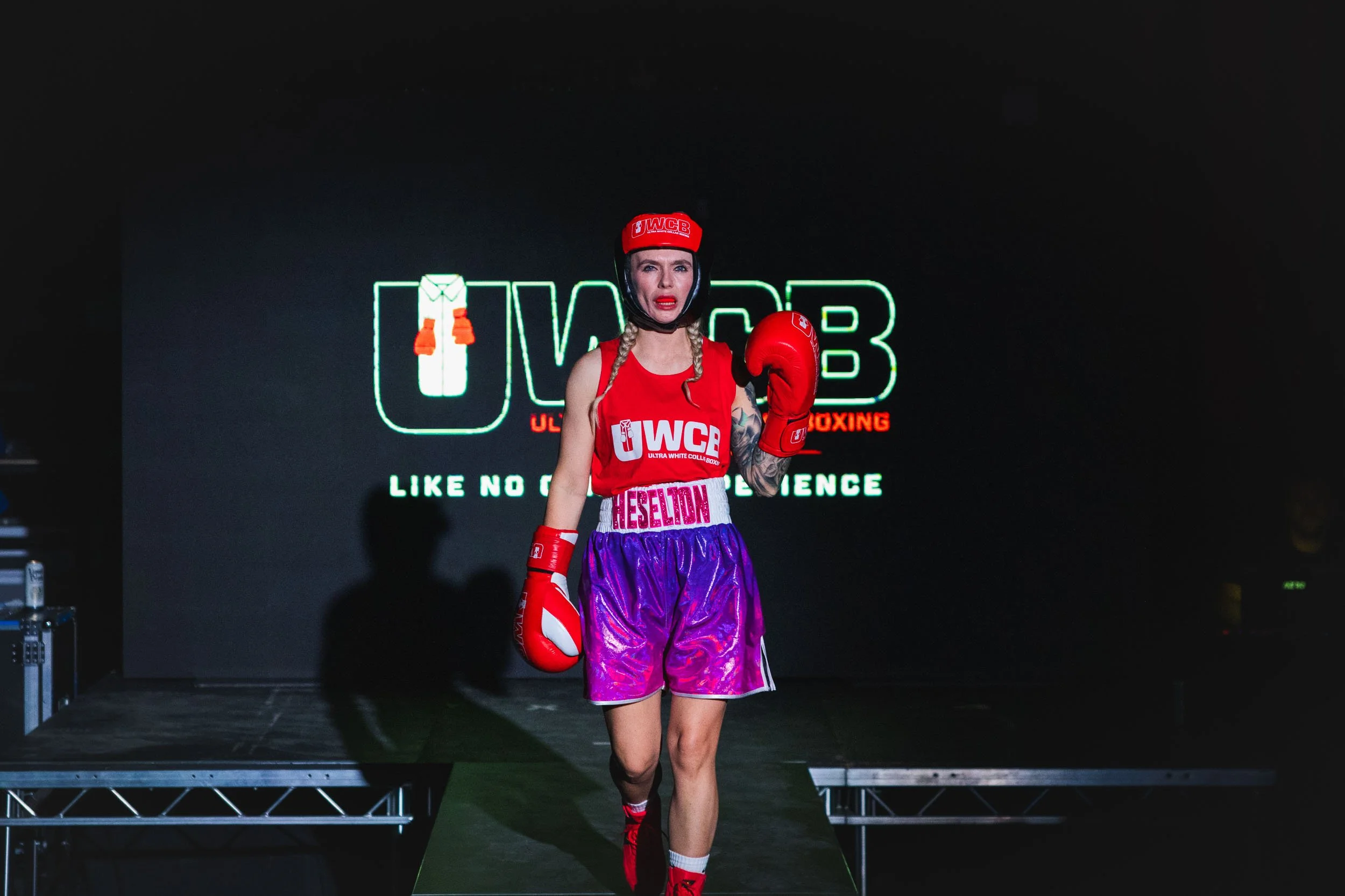 Female boxer walking on the ring, wearing red boxing gloves, red headgear, purple shorts, and a red tank top with UFC branding. She has blonde hair in braids and tattoos on her left arm.