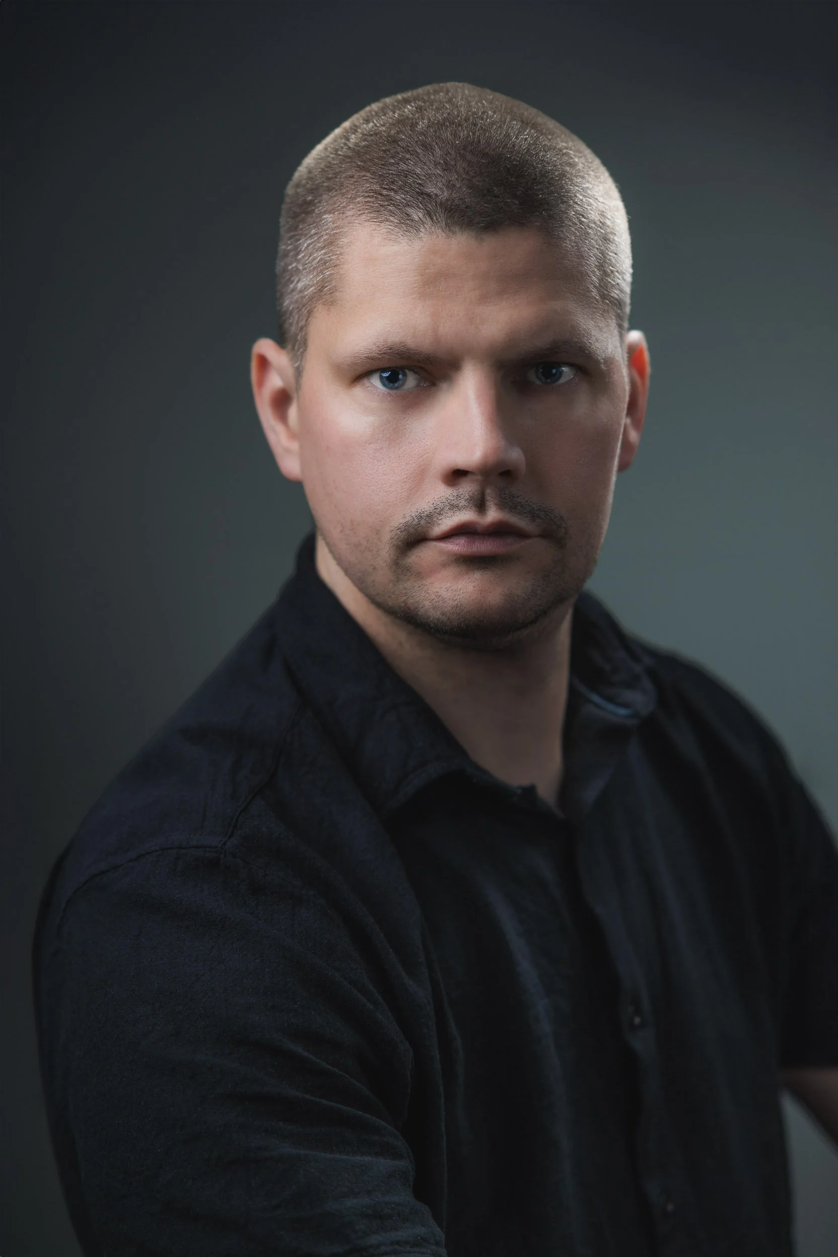Portrait of a man with short light brown hair, blue eyes, and fair skin, wearing a dark shirt against a dark background.