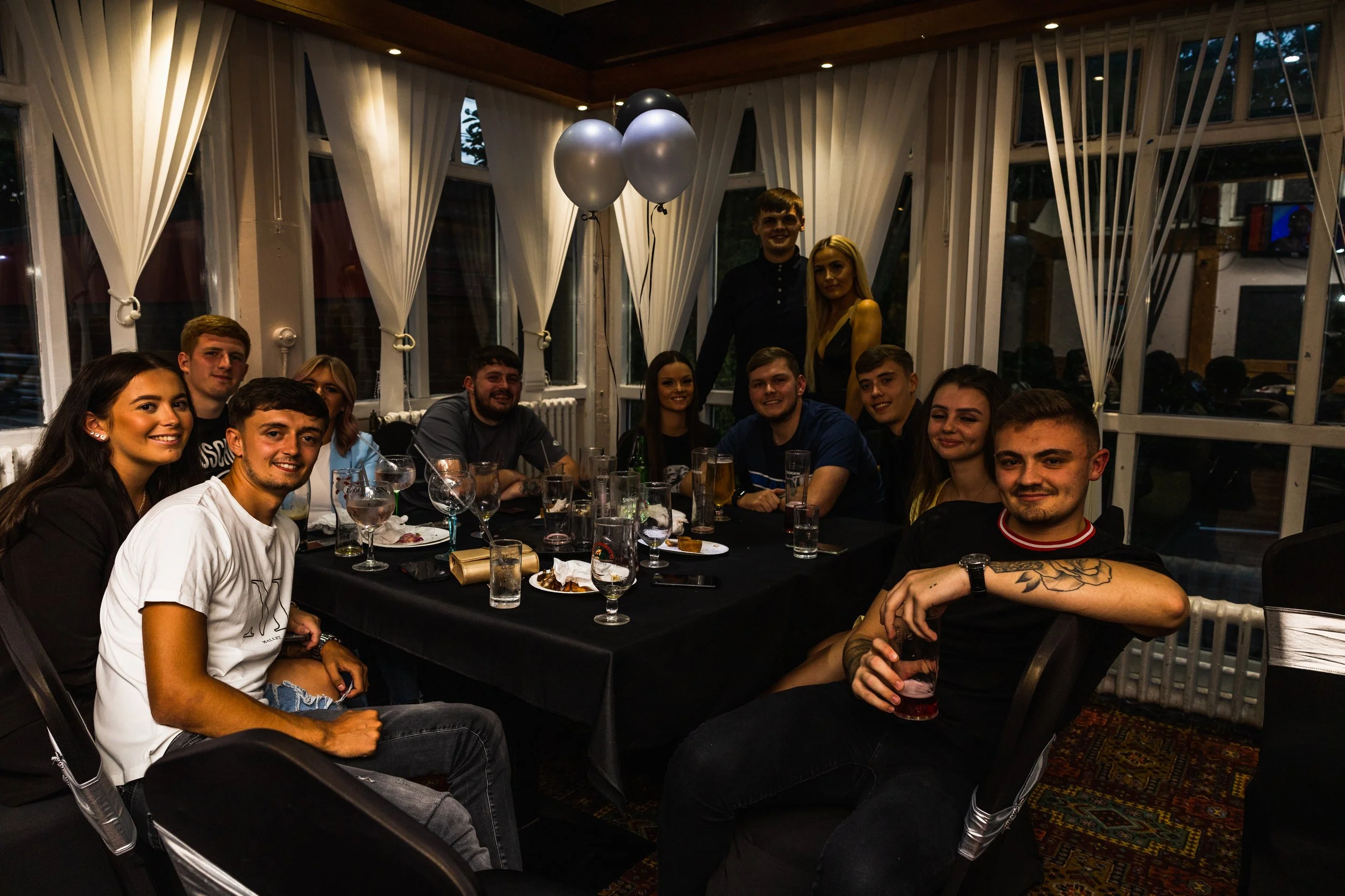A group of young adults gathered around a table at a celebration, with balloons and drinks in a decorated indoor setting.