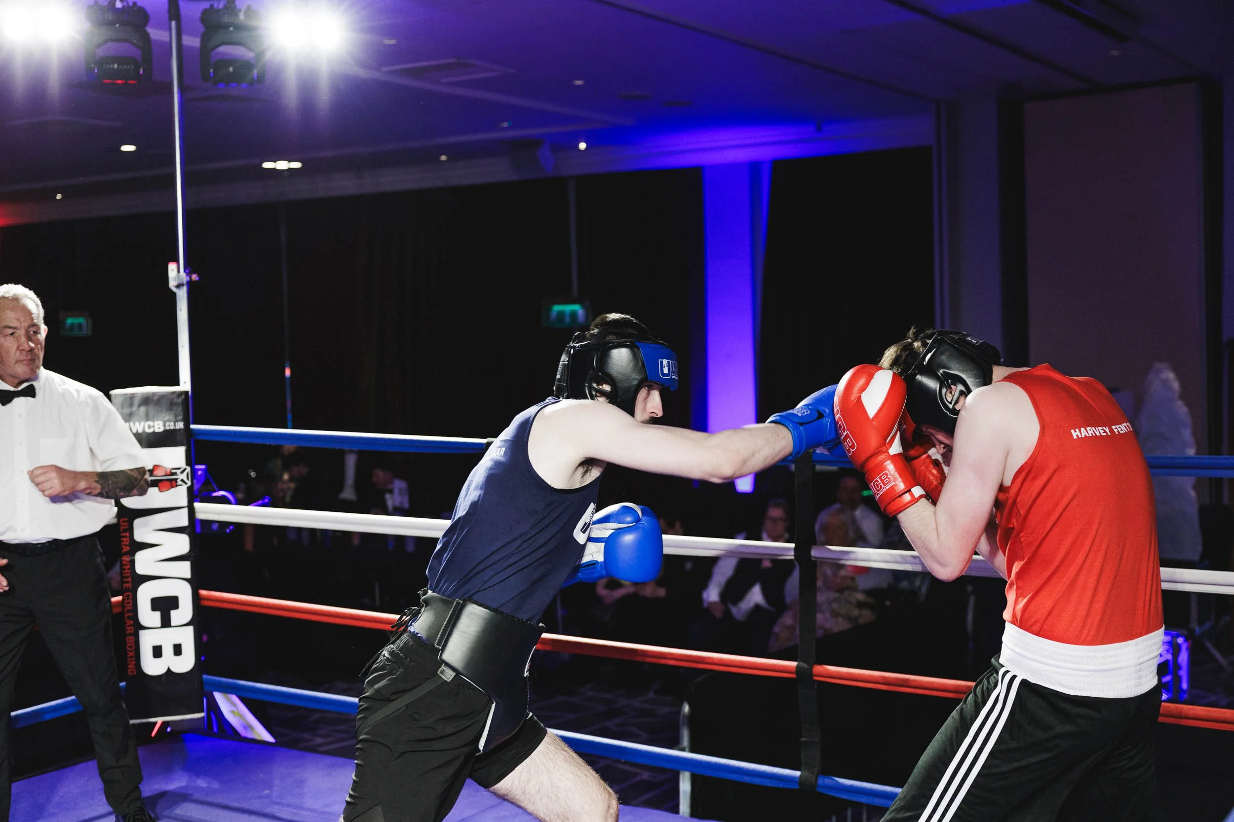 Two amateur boxers, one in a blue shirt and the other in a red shirt, are inside a boxing ring engaged in a match. The boxer in blue is throwing a punch while the boxer in red is covering his face with gloves. An official dressed in a white shirt and