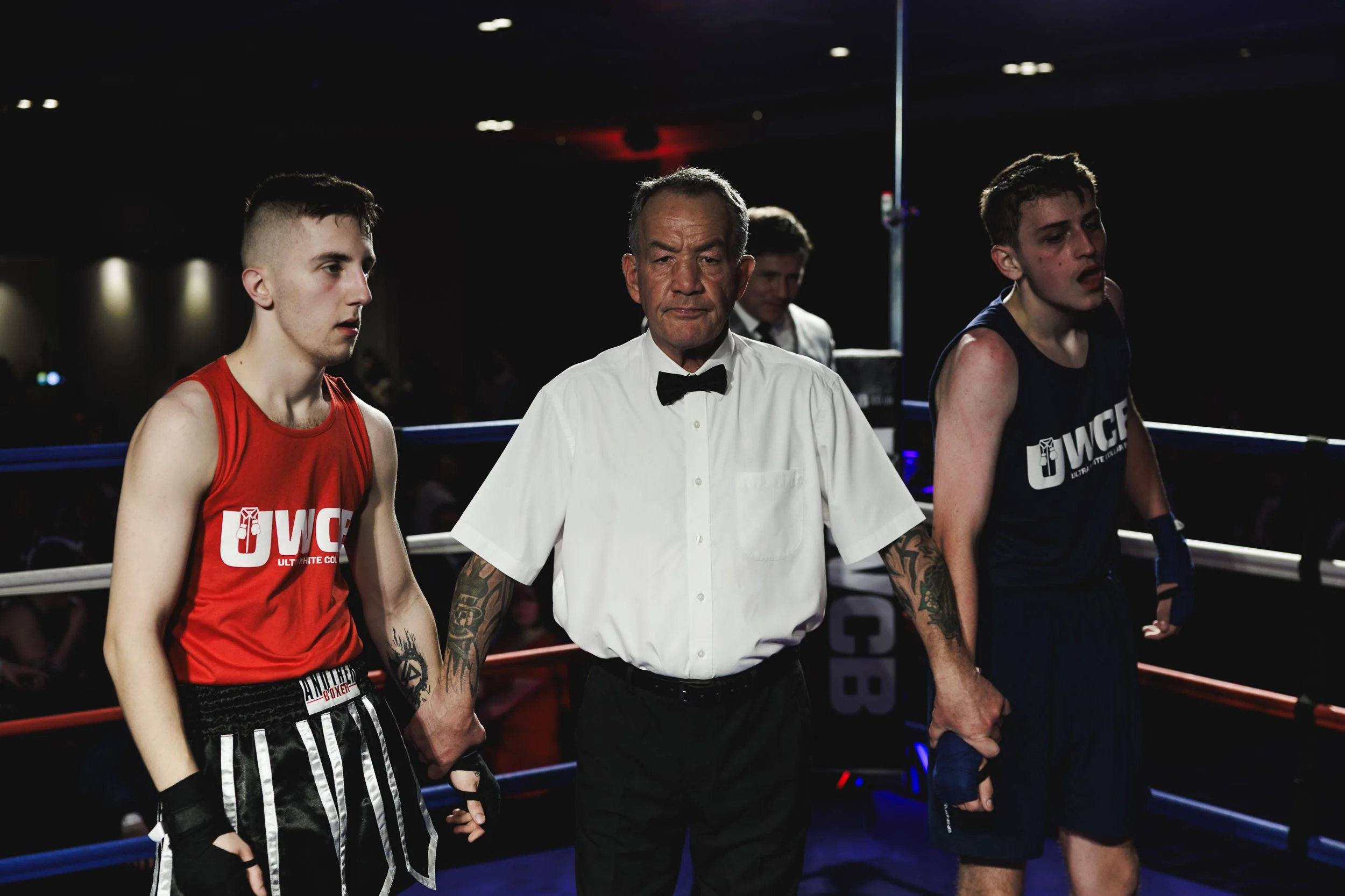 Two young male boxers in boxing ring with referee between them, holding hands after match. Left boxer wears red shirt and black shorts, right boxer wears black shirt and shorts. The scene appears intense with dark background and audience.