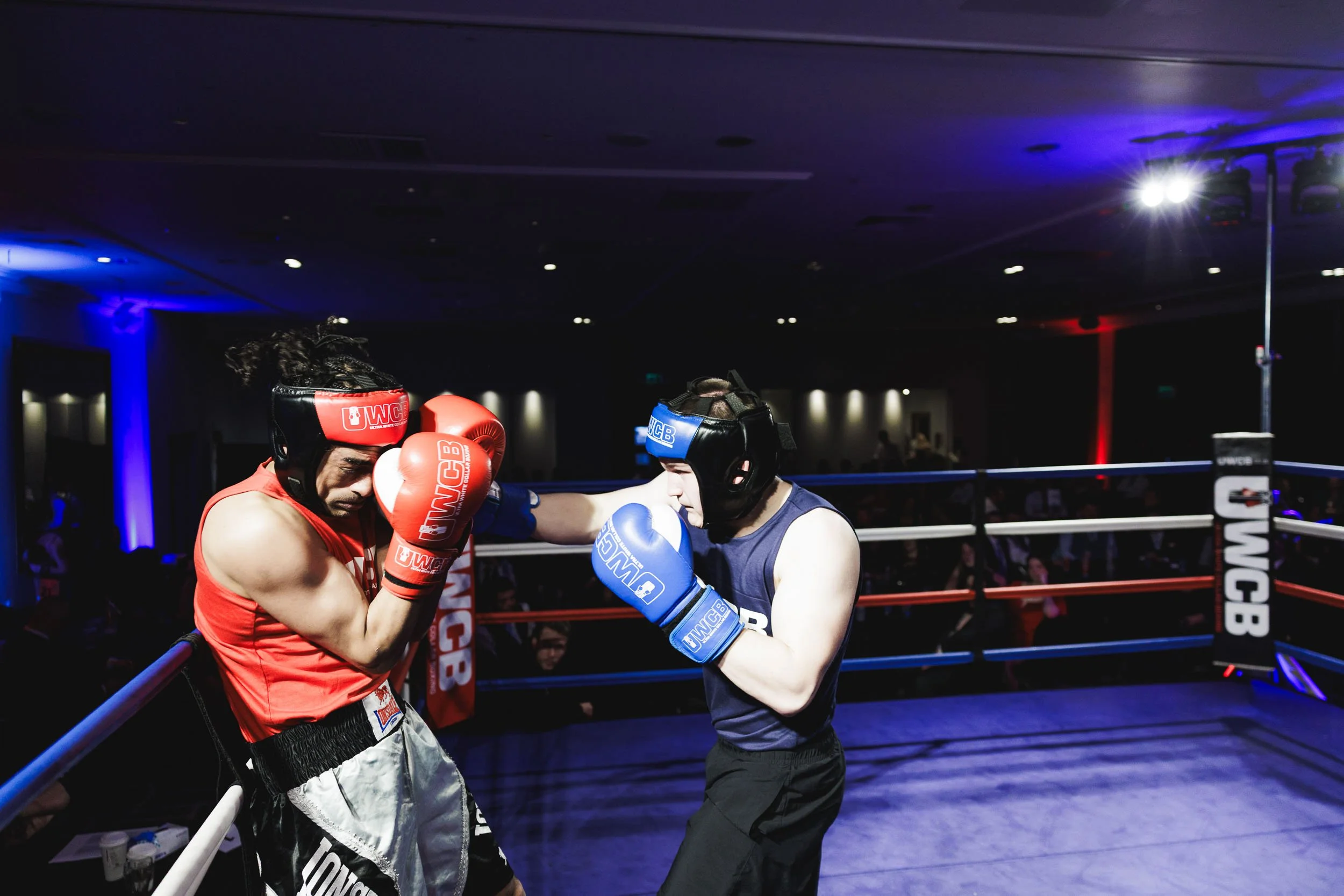 Two male boxers wearing protective headgear and gloves sparring inside a boxing ring, with one in red gear and the other in blue gear.