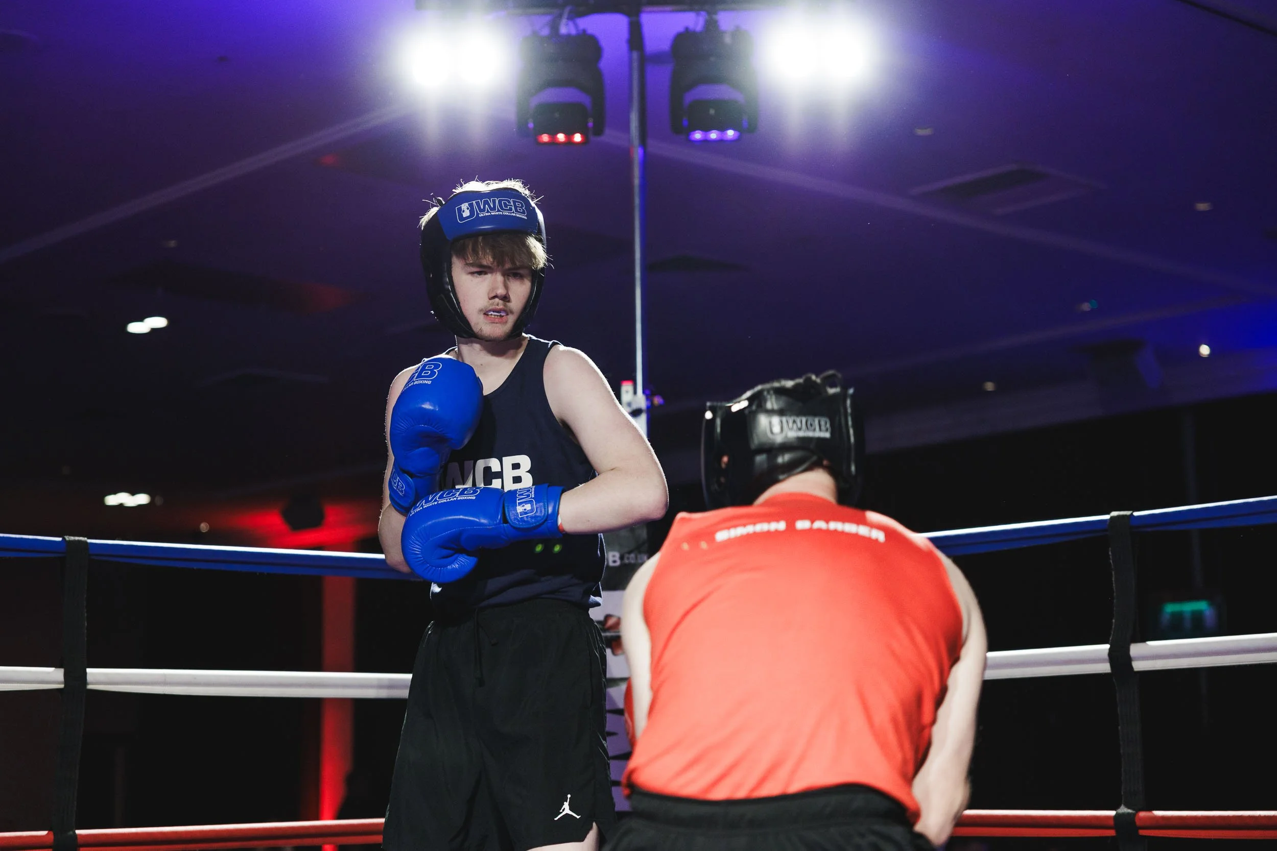 A young male boxer wearing black shorts and a sleeveless top with boxing gloves and a helmet stands in a boxing ring, looking at his opponent, who is crouched and wearing orange and black gear.