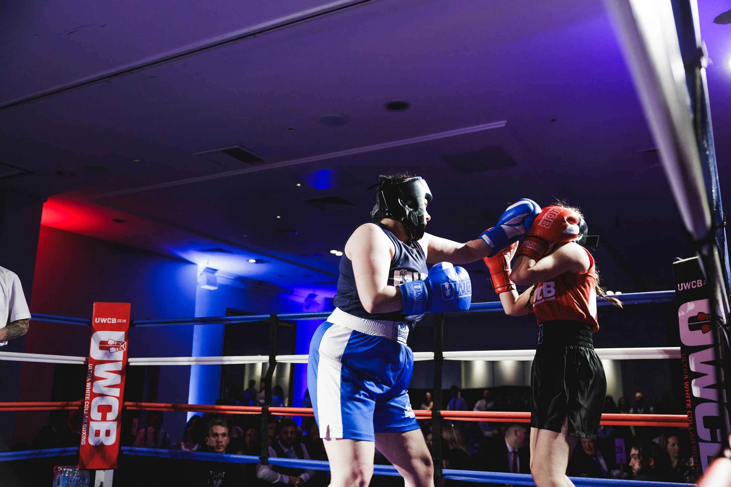 Two women engaged in a boxing match inside a ring with white, red, and blue ropes. One woman in blue shorts and top is landing a punch on the woman in red shorts and top. Both women wear protective headgear and boxing gloves. Audience members are vis