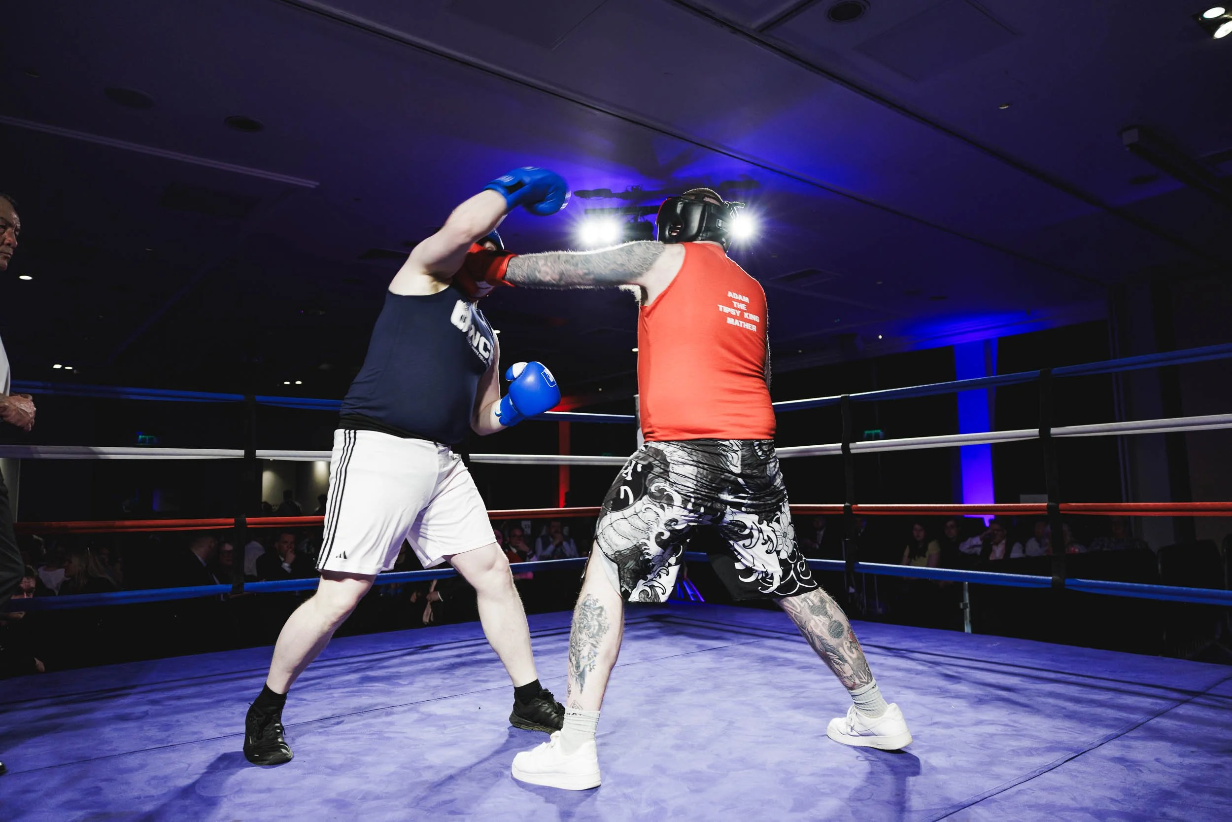 Two boxers in a ring sparring, one in black gloves and T-shirt, the other in blue gloves and shorts, throwing punches at each other. An audience watches in the background.