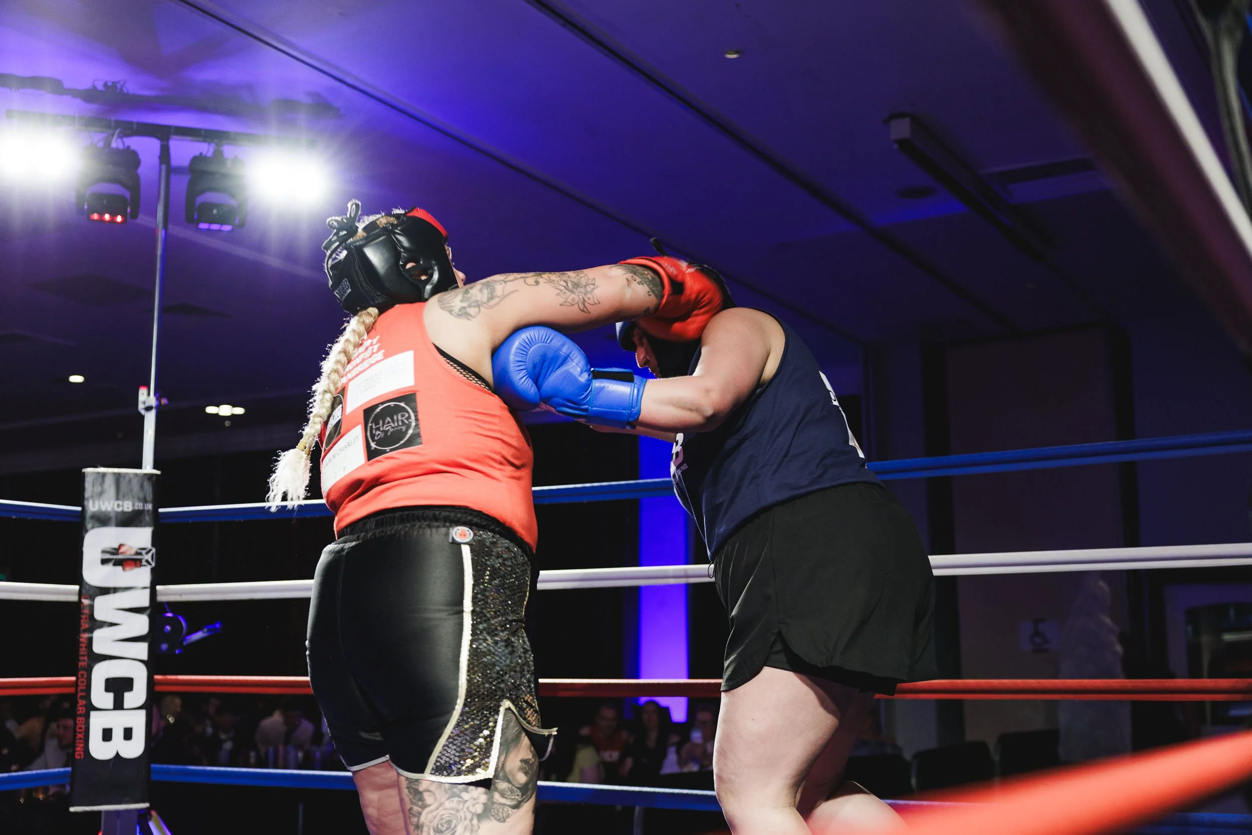 Two female boxers wearing protective headgear and gloves sparring in a boxing ring, with one delivering a punch to the other's face. Bright lights shine down on the ring.