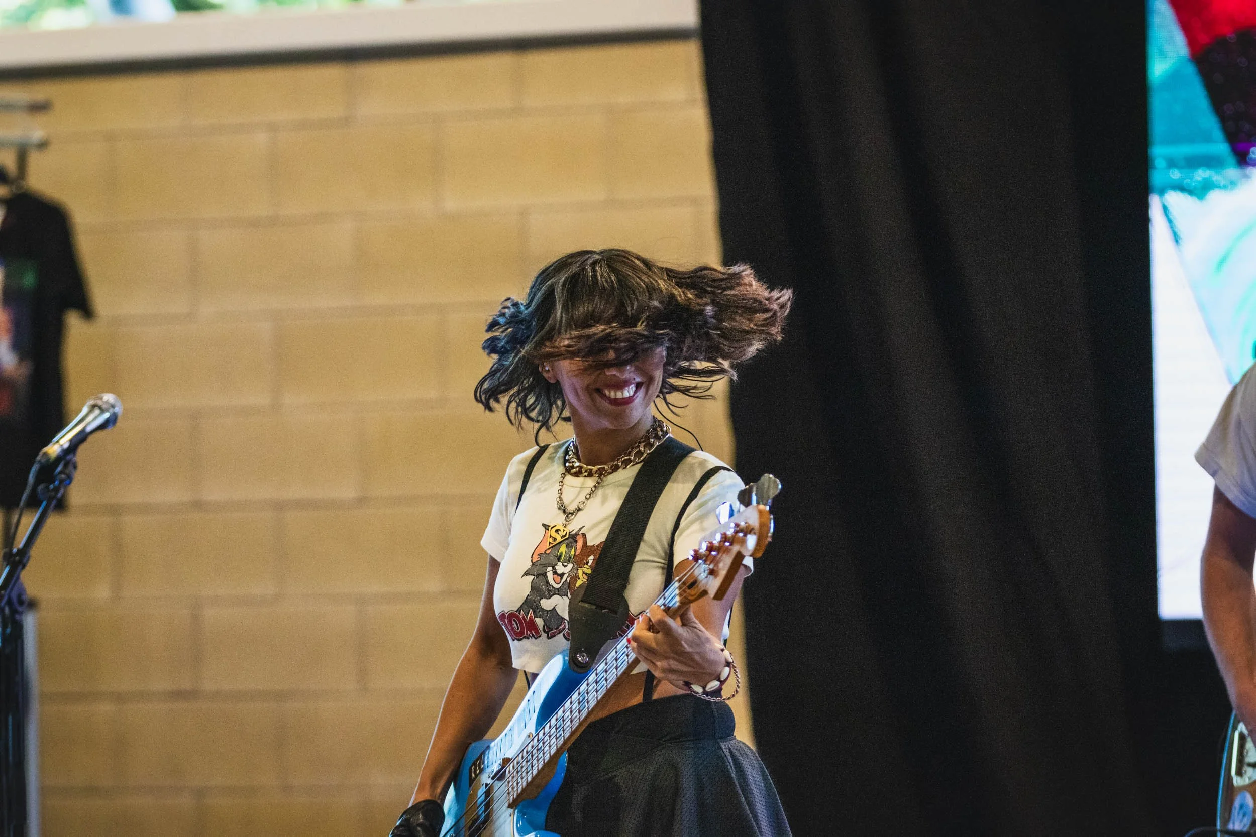A woman with dark hair playing an electric guitar and smiling, wearing a graphic t-shirt, necklace, and skirt, in a room with a beige brick wall and black curtain.