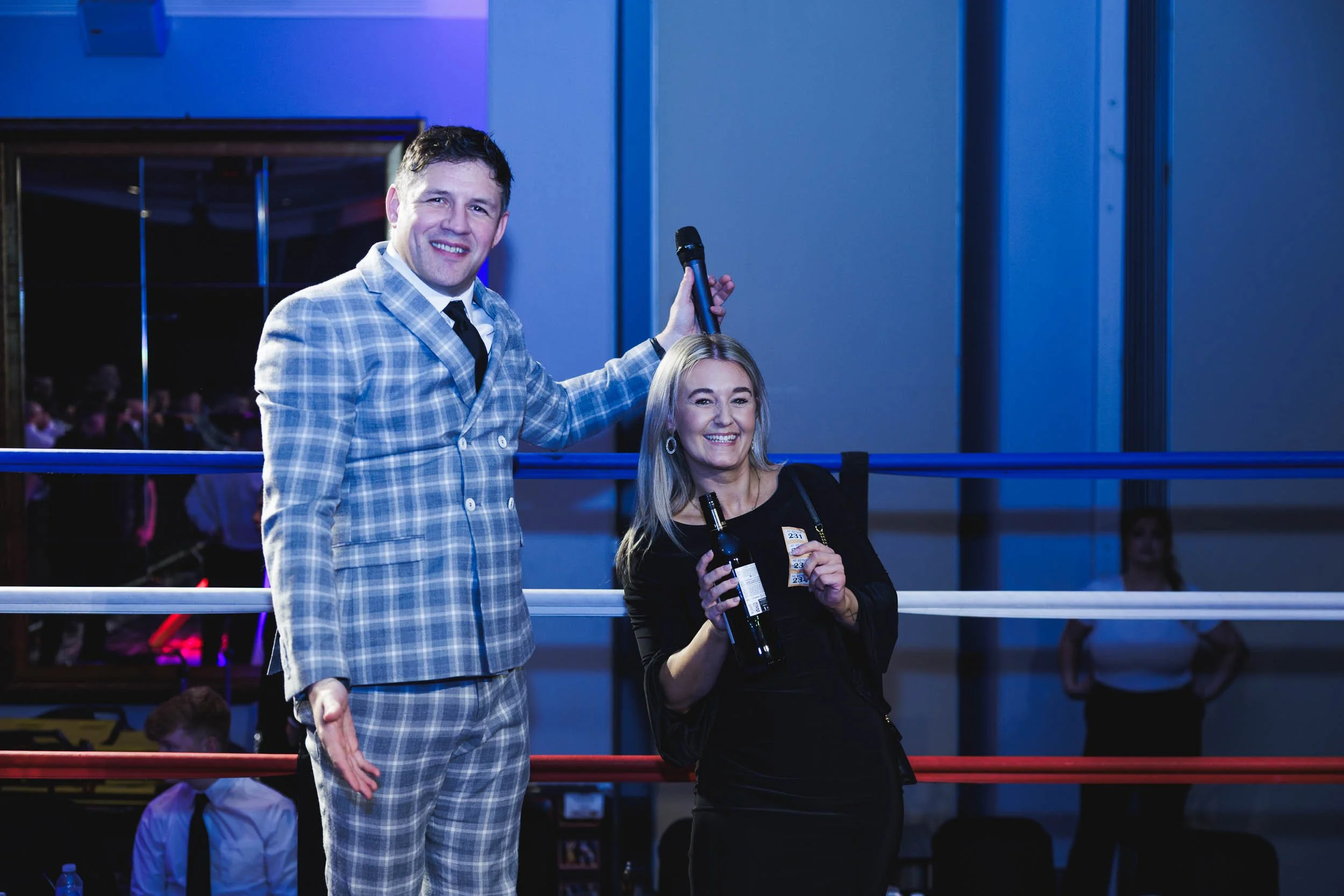 Man in plaid suit and woman in black dress standing inside a boxing ring, smiling and holding bottles of alcohol, with blue and red ropes behind them.