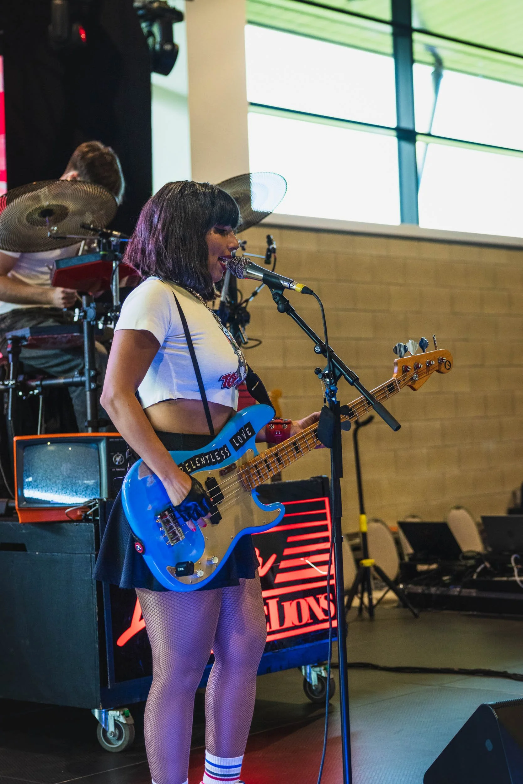 A woman singing into a microphone while playing a blue electric bass guitar during a live performance, with a drummer in the background
