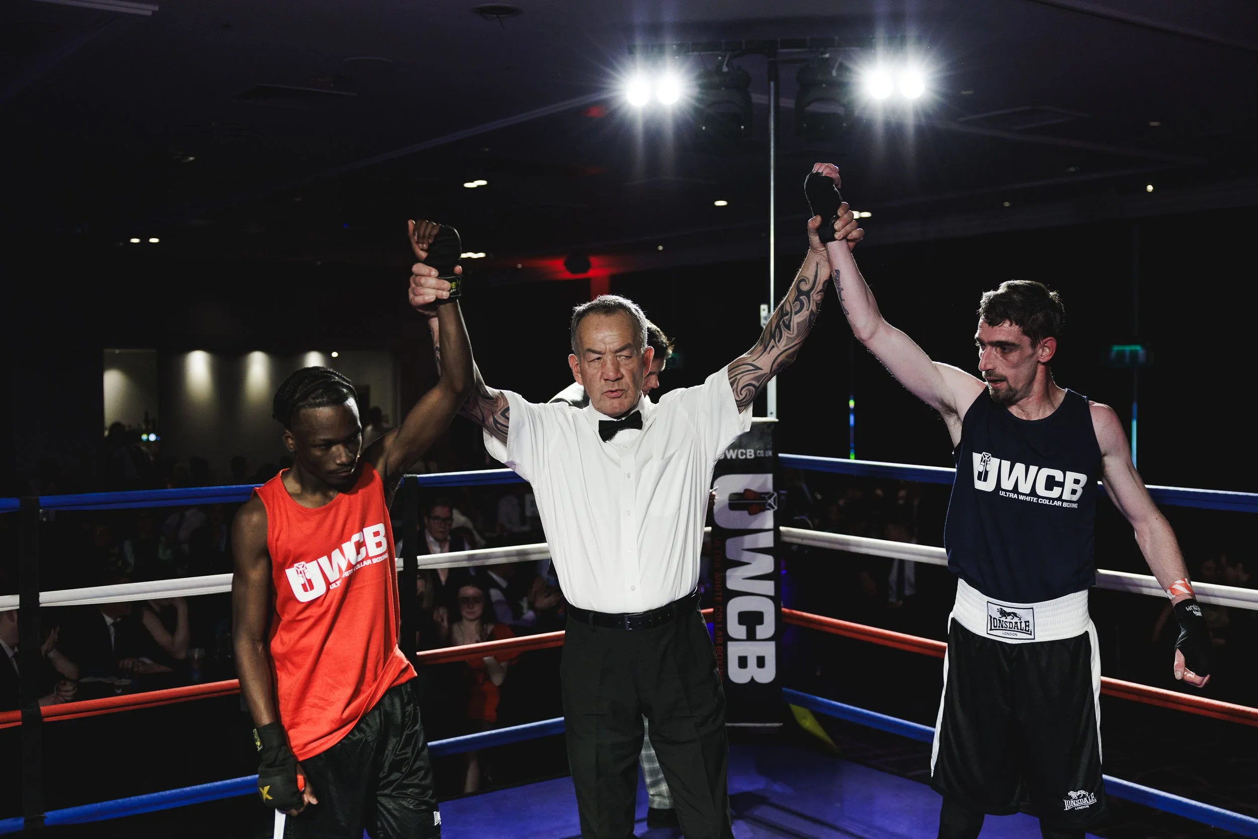 Boxing match ending with a referee declaring the winner. Two boxers with raised arms: one in a red tank top and the other in a black tank top, inside a boxing ring at a professional event.