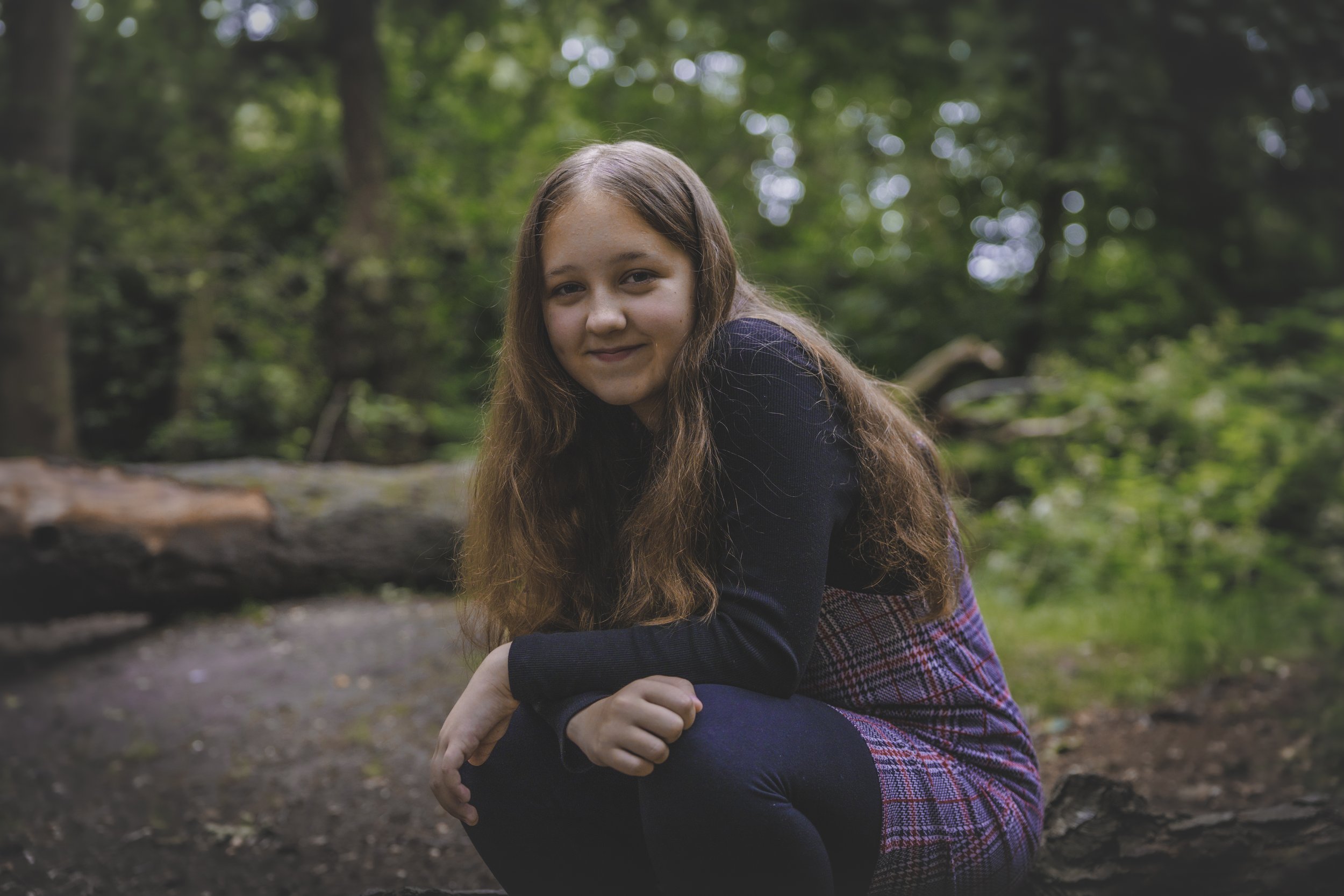A young girl with long brown hair smiling at the camera while sitting outdoors in a wooded area with green trees and a fallen log in the background.