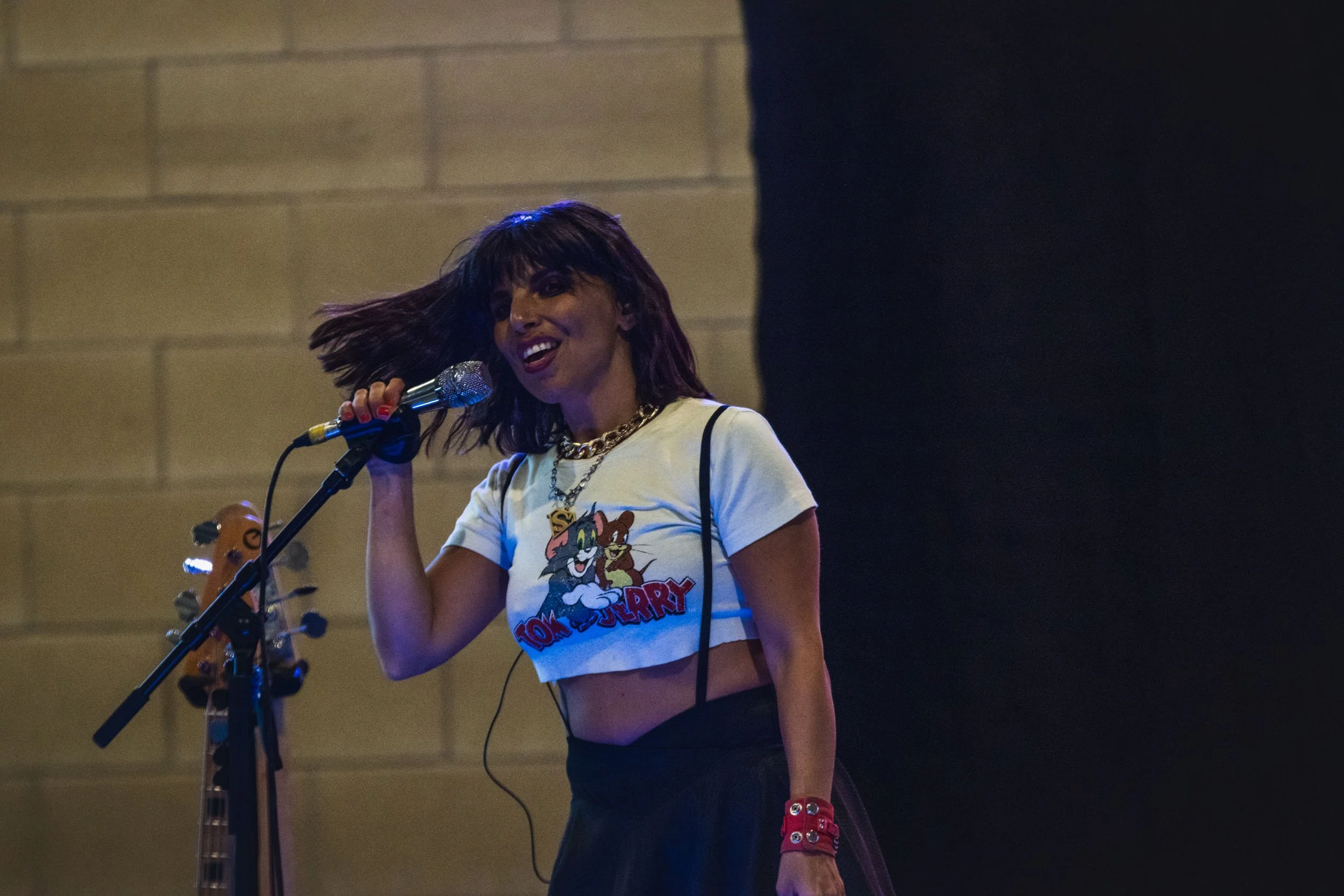 A woman singing into a microphone on stage, wearing a Tom and Jerry crop top, a black skirt, and red accessories, with a guitar in the background.