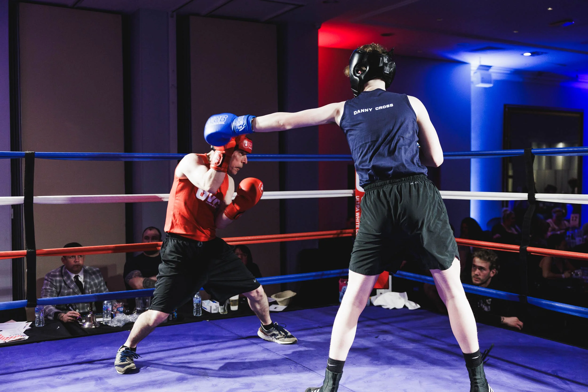 Two men boxing in a ring. The man on the left wearing red gloves and gear is throwing a punch, while the man on the right wearing black shorts and gloves is blocking or countering. Spectators seated around the ring watch the match.