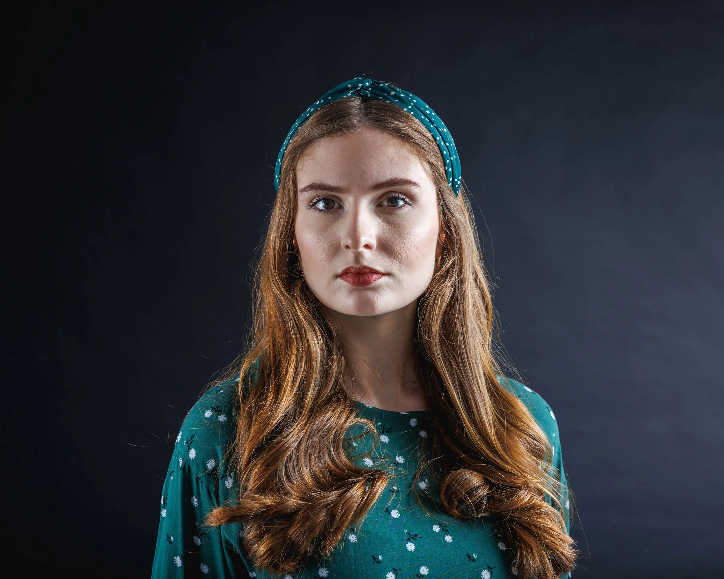 Portrait of a young woman with long wavy red hair, wearing a teal headband with white polka dots and a matching teal top with white floral pattern, against a dark background.