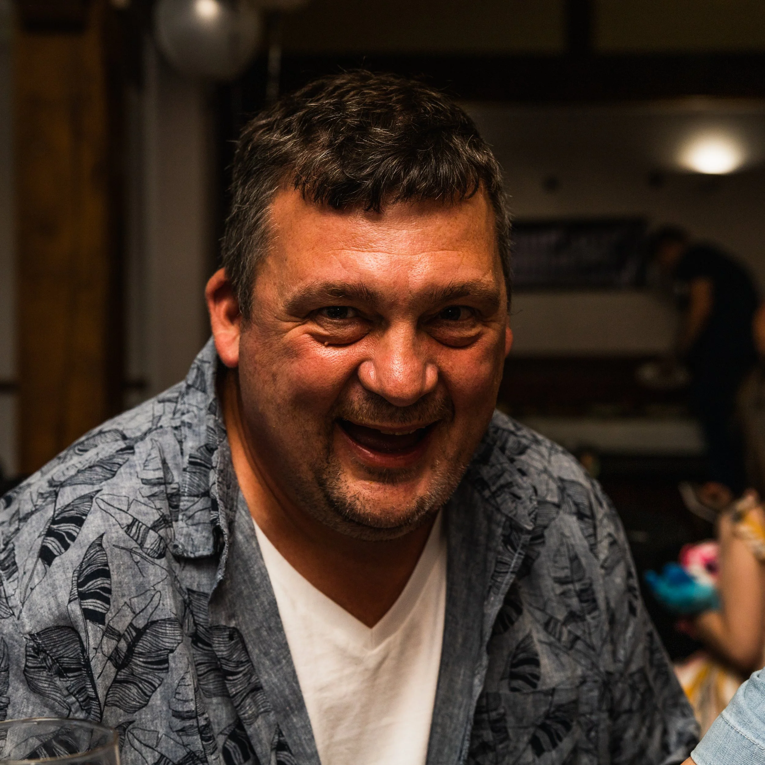 A middle-aged man with short dark hair and a beard, smiling and laughing indoors at a social gathering.