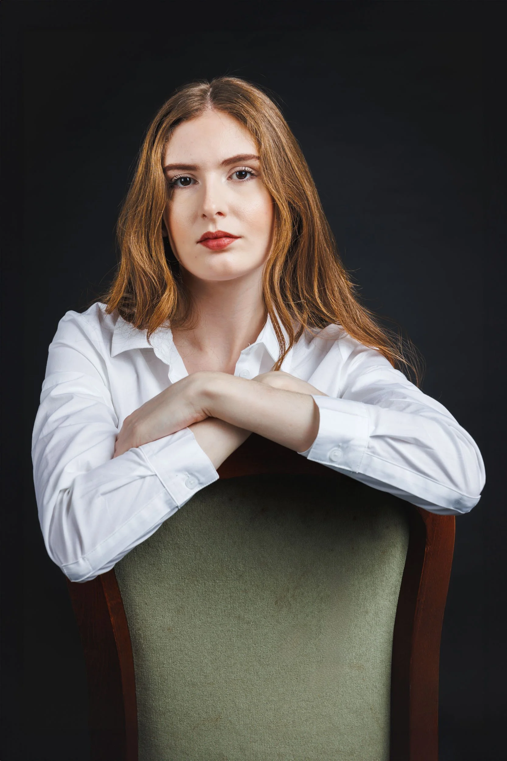 Portrait of a young woman with reddish-brown hair and fair skin, seated with arms crossed on the back of a green chair, wearing a white button-down shirt against a dark background.