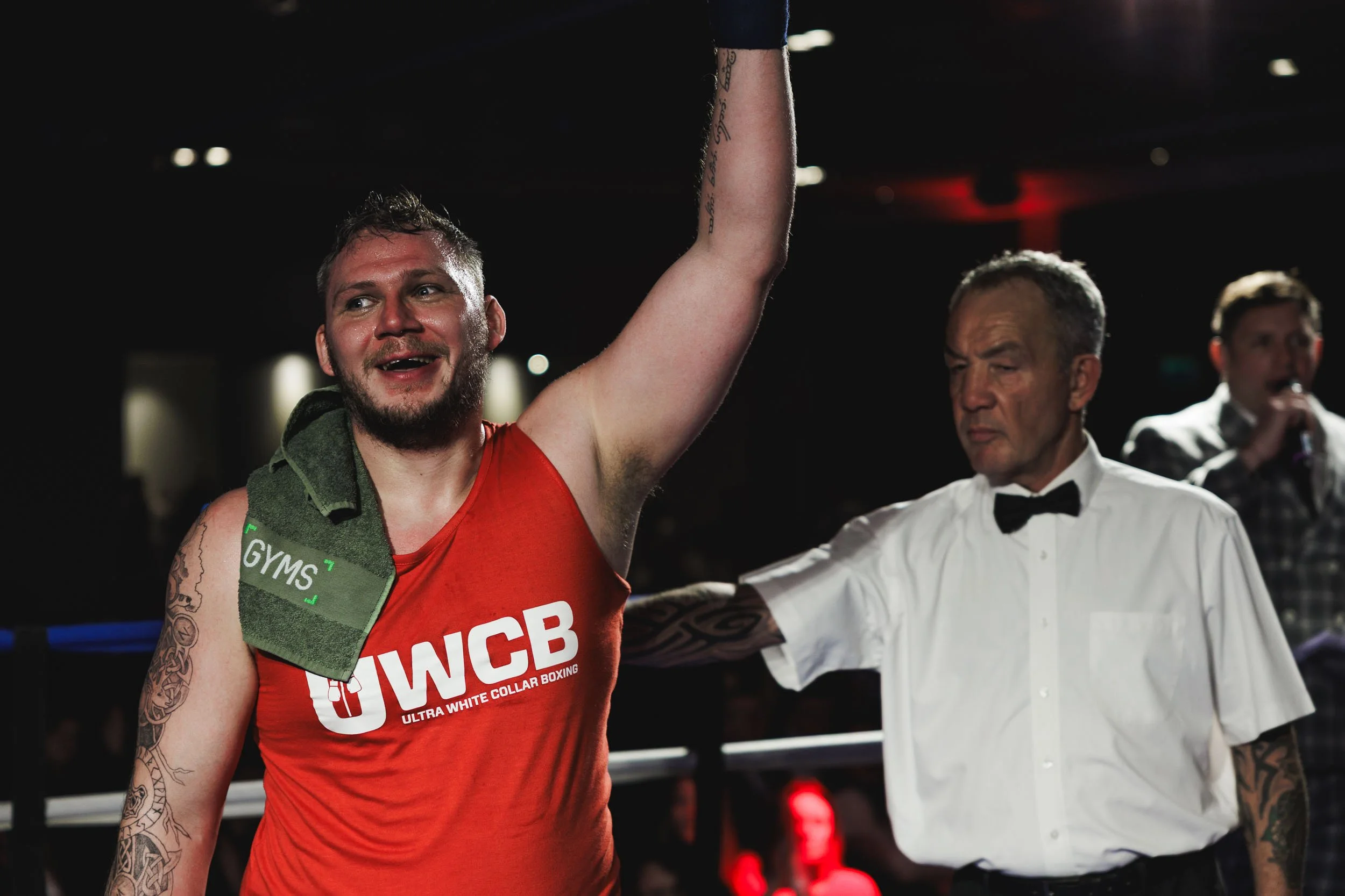 A male boxer with a towel draped over his shoulder, wearing a red shirt with 'UWCB' and 'Ultra White Collar Boxing' on it, raises his arm in victory inside a boxing ring. An official or referee wearing a white shirt with a black bow tie is touching h