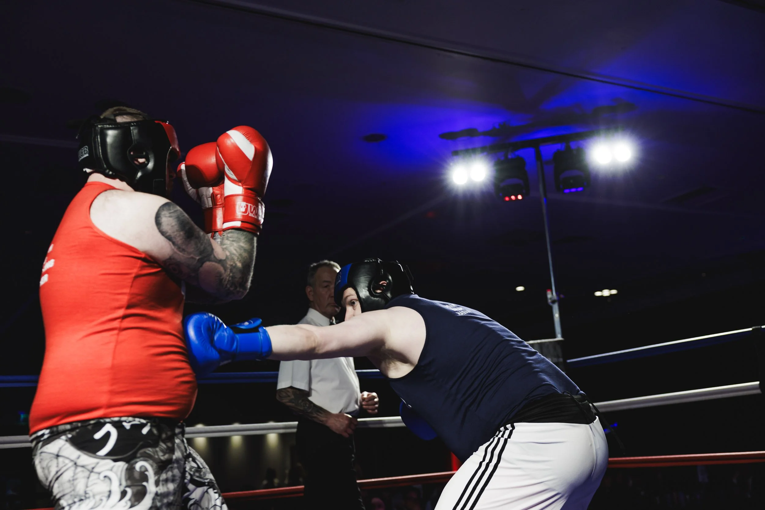 Two men sparring in a boxing ring, wearing protective headgear and gloves, with a referee observing in the background under bright lights.