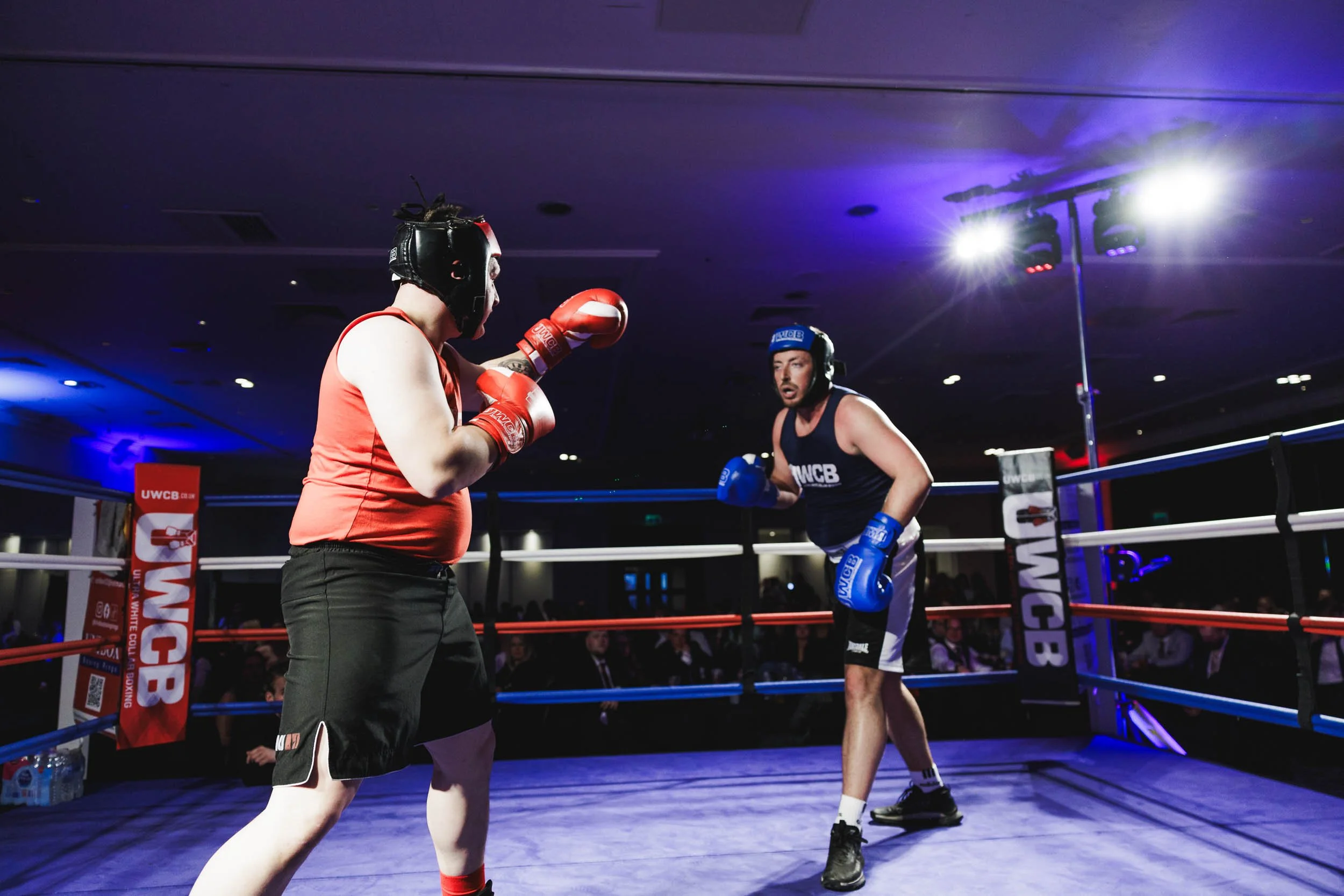 Two men in boxing gear sparring in a boxing ring, with audience in the background and bright lights overhead.