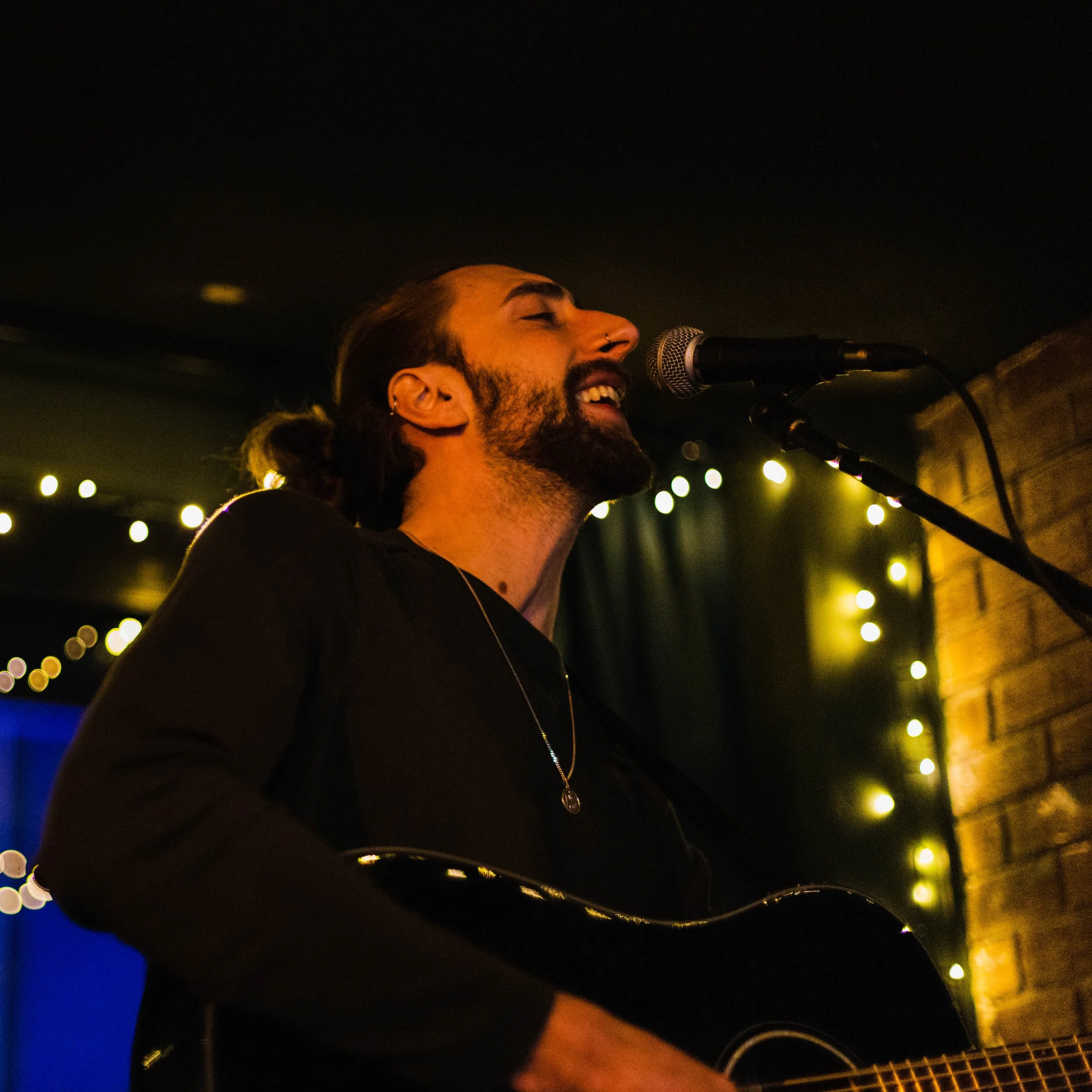 A man with a beard and long hair in a ponytail is singing into a microphone while playing an acoustic guitar. He is smiling with his eyes closed, in a dimly lit setting with string lights in the background.