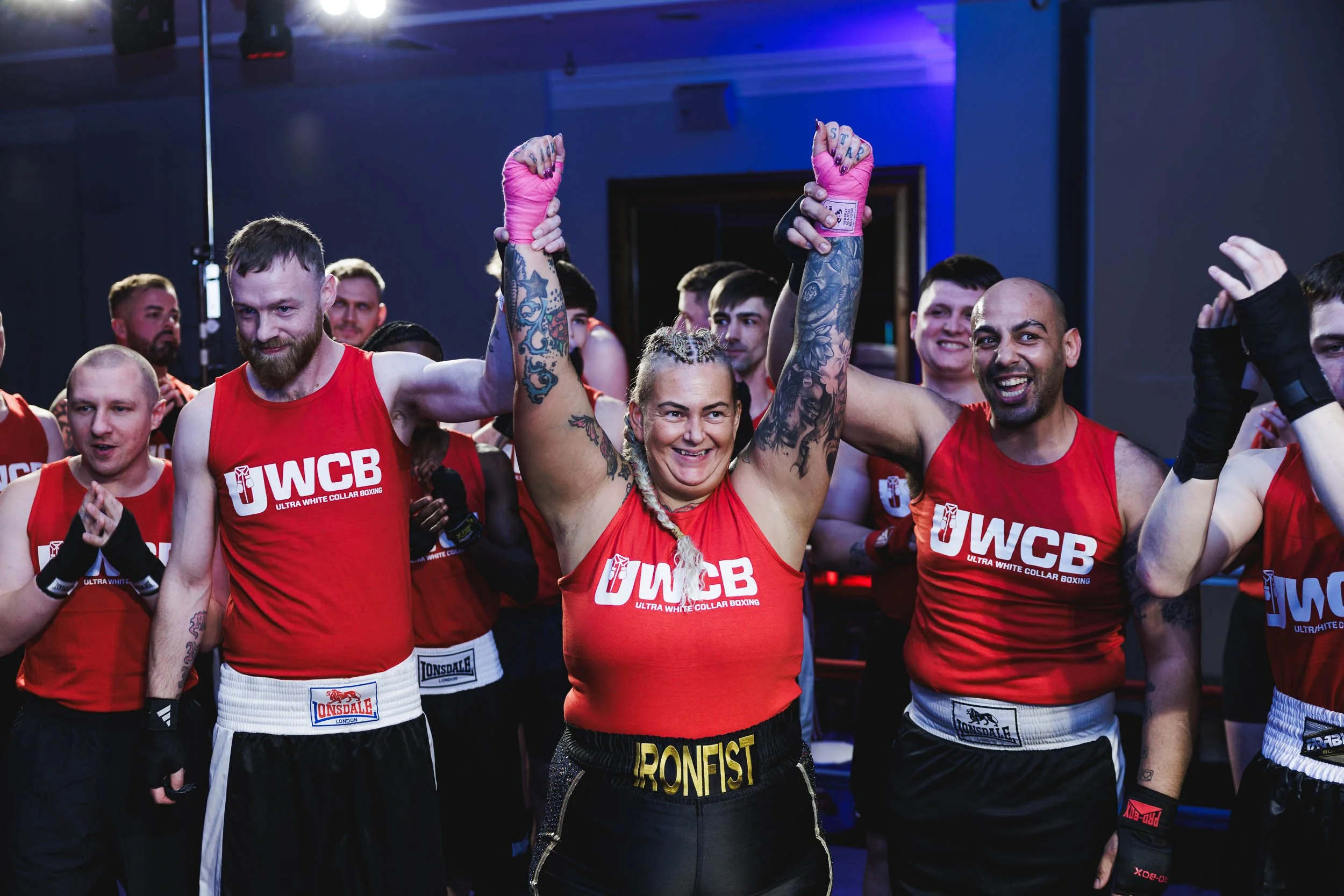 Boxers celebrating after a match, standing in a gym, wearing red shirts with 'UWCB' logos and black shorts, raising fists in victory, surrounded by fellow fighters clapping and smiling.