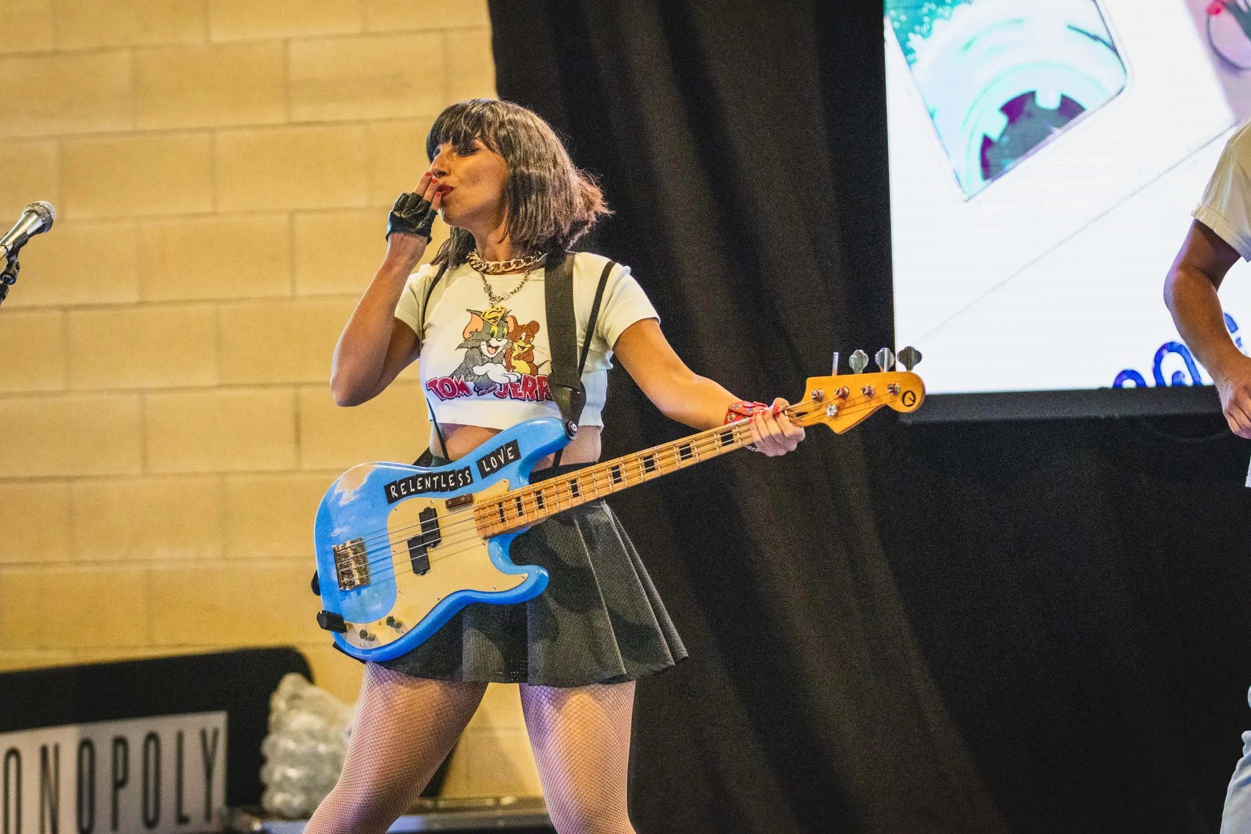 A woman in a Tom and Jerry crop top and a black skirt holding a blue bass guitar with stickers that say 'Relentless Love' while performing on stage.