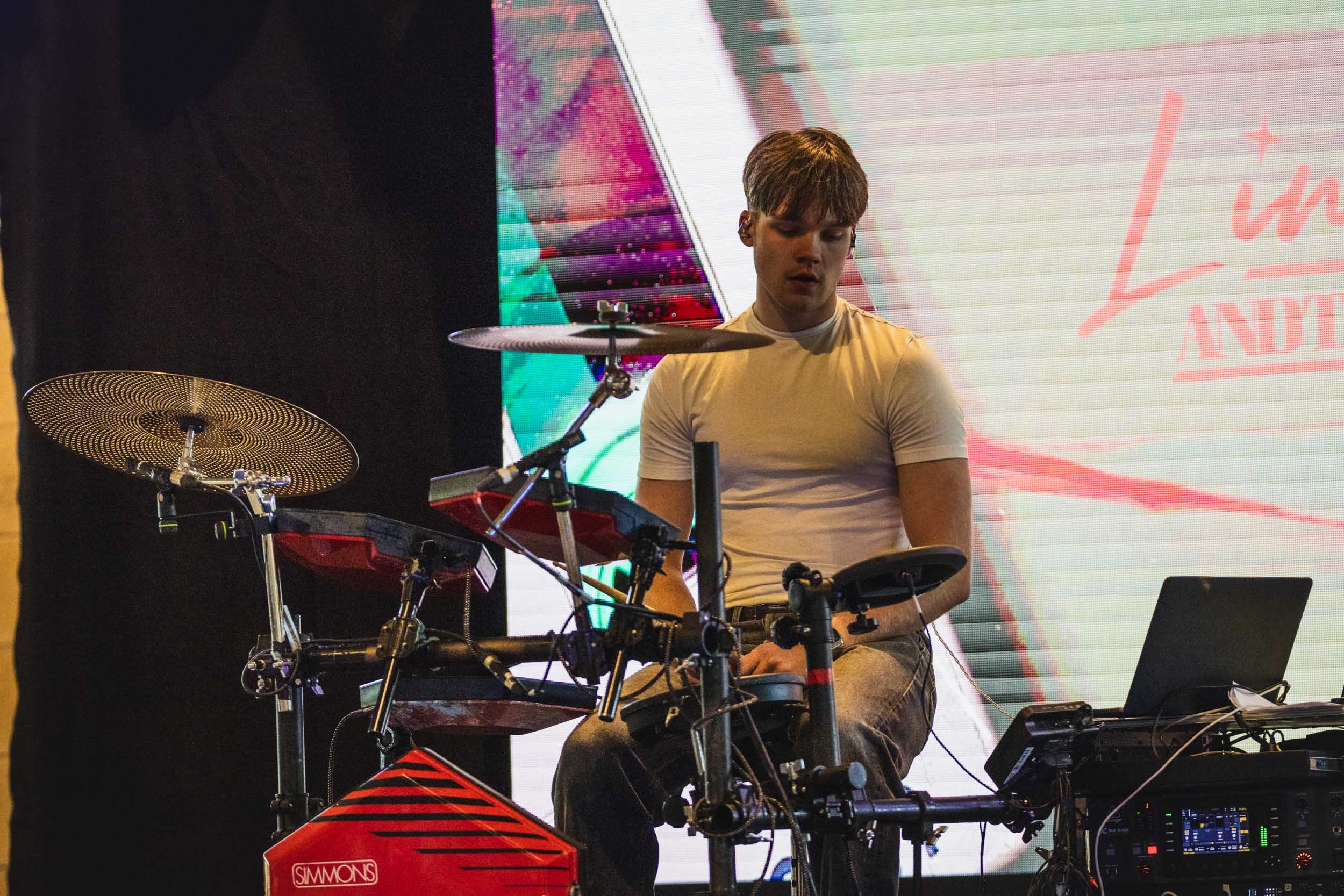 A young man playing a drum pad set on stage, with electronic equipment and a large colorful screen in the background.