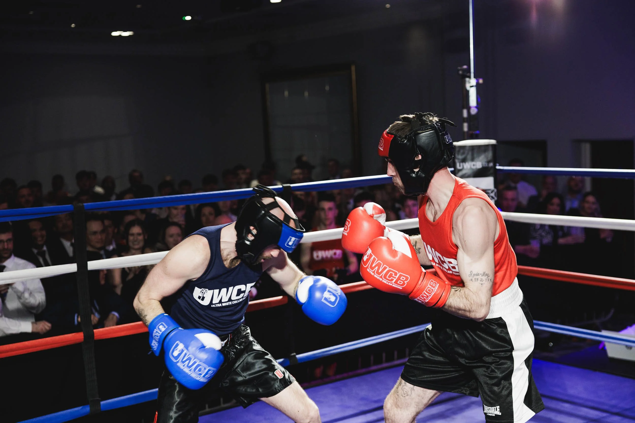 Two male boxers wearing protective headgear and boxing gloves sparring in a ring, with an audience watching in the background.
