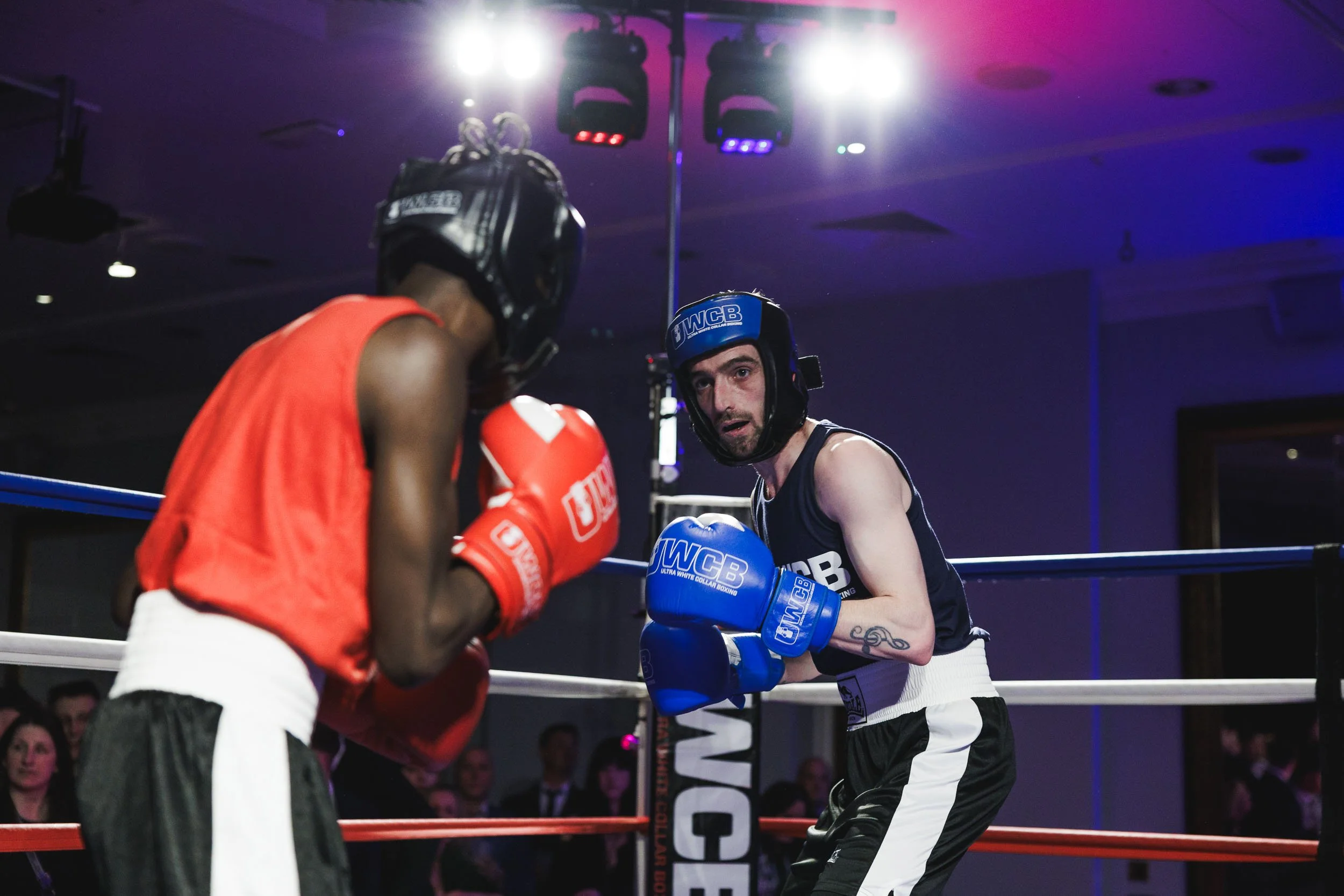 Two male boxers in a boxing ring, wearing protective headgear, boxing gloves, shorts, and tank tops, engaging in a match under colorful lighting with audience members watching in the background.