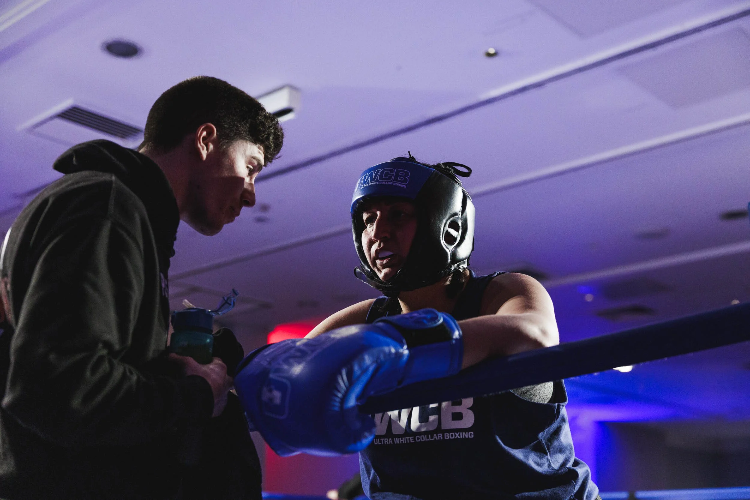 A female boxer with a protective helmet and blue boxing gloves resting on the corner of the ring, listening to her coach during a match or training, with a male coach talking to her, in an indoor boxing gym.