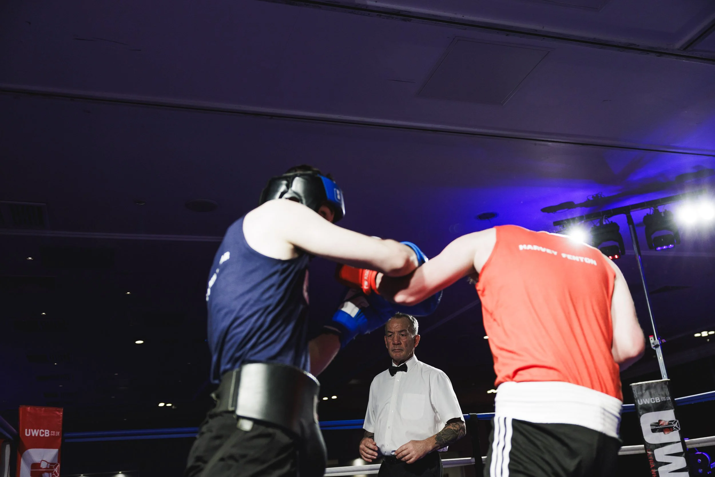 Two male boxers wearing protective headgear and gloves sparring in a boxing ring, with a referee watching closely in the background.