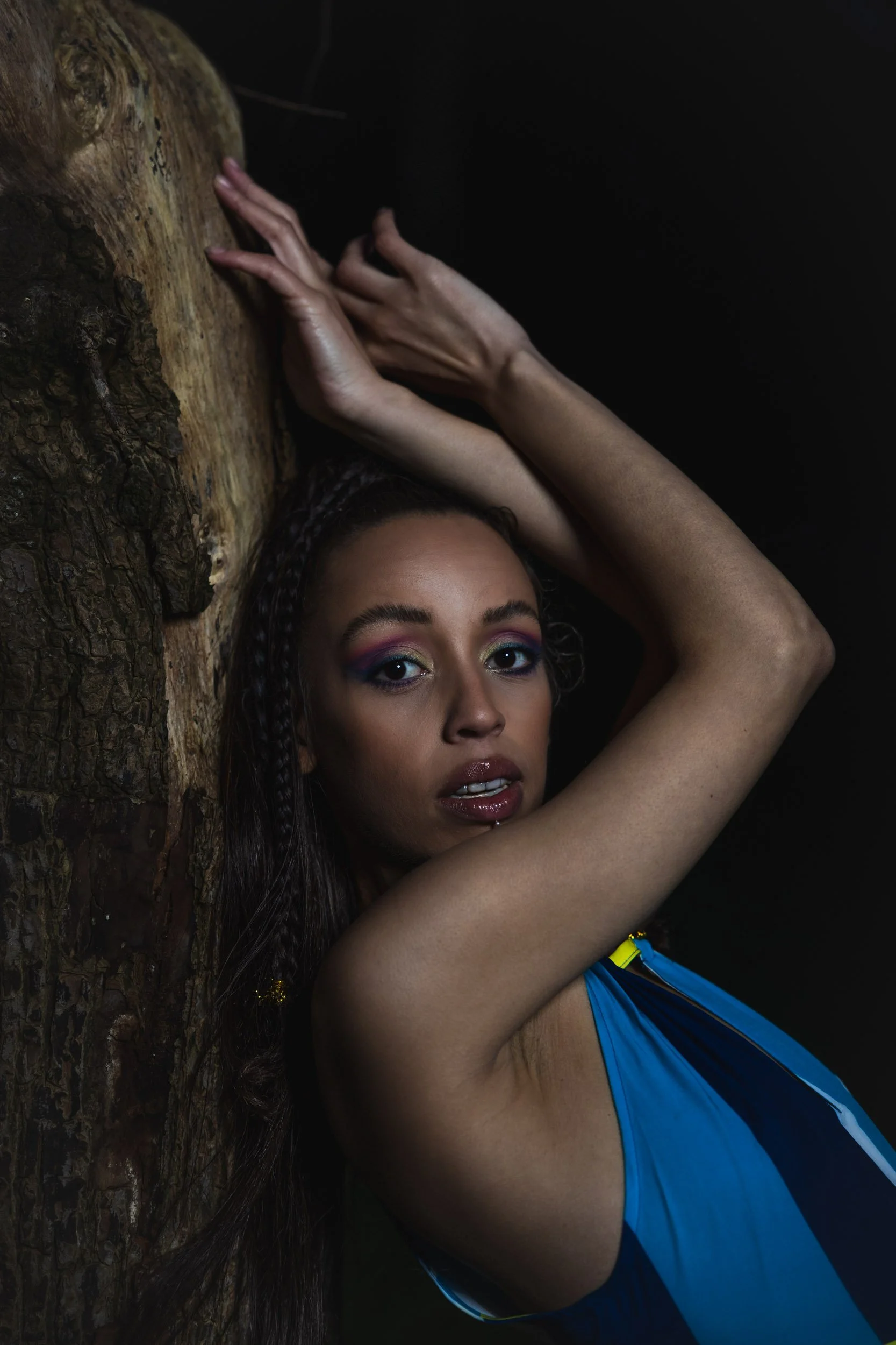 A woman with colorful eye makeup and dark hair styled in braids leans against a textured tree trunk with her arms raised, wearing a blue sleeveless top.