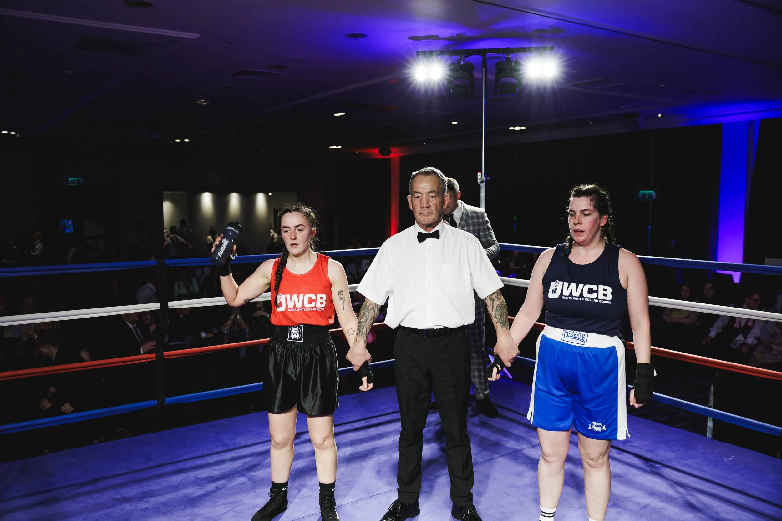 Two female amateur boxers in boxing gloves and sports gear standing in a boxing ring, holding hands with a referee after a match, with spectators in the background.