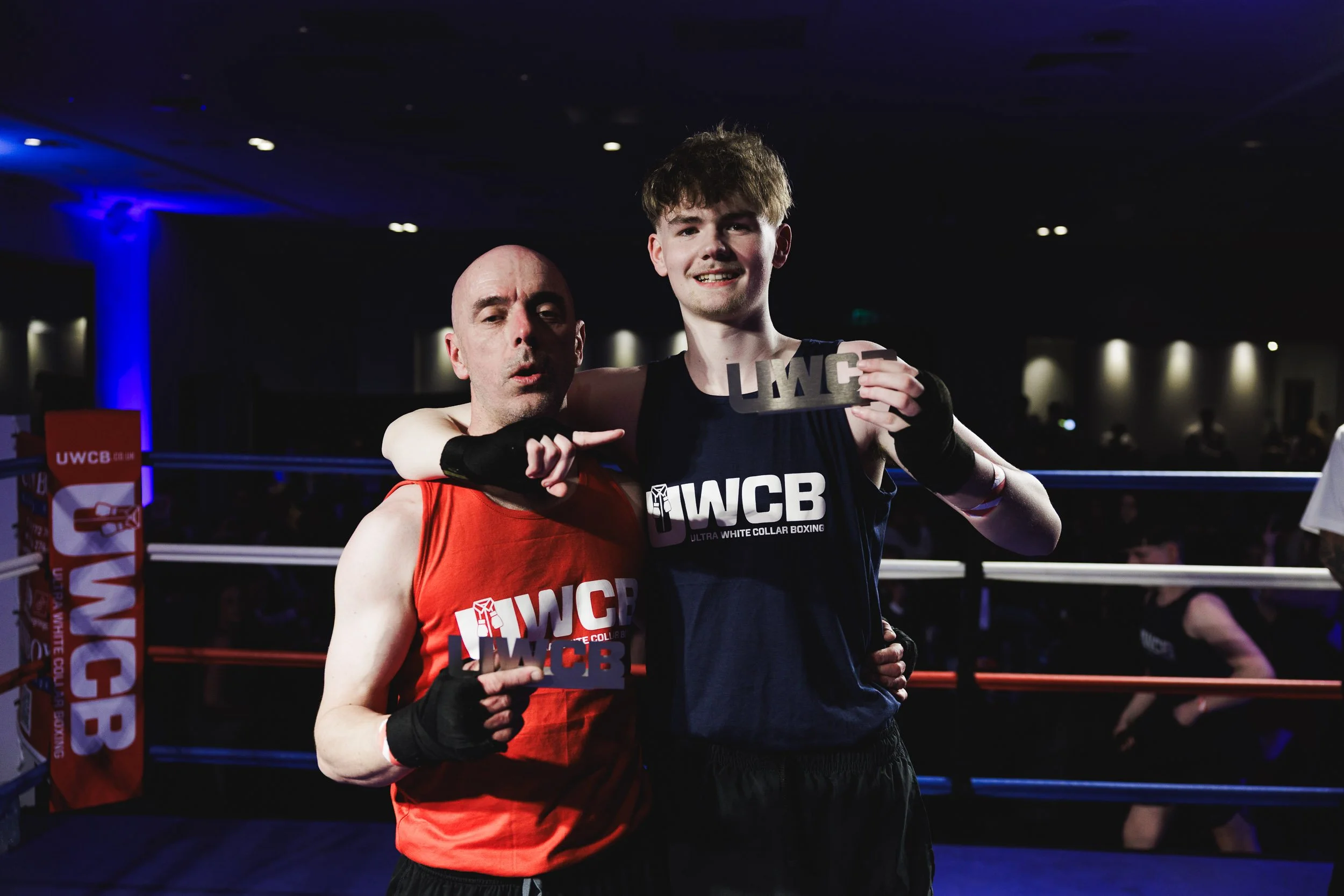 Two male boxers pose inside a boxing ring, celebrating a match. One boxer is wearing a red tank top, and the other is wearing a navy blue tank top. They are holding up prizes with