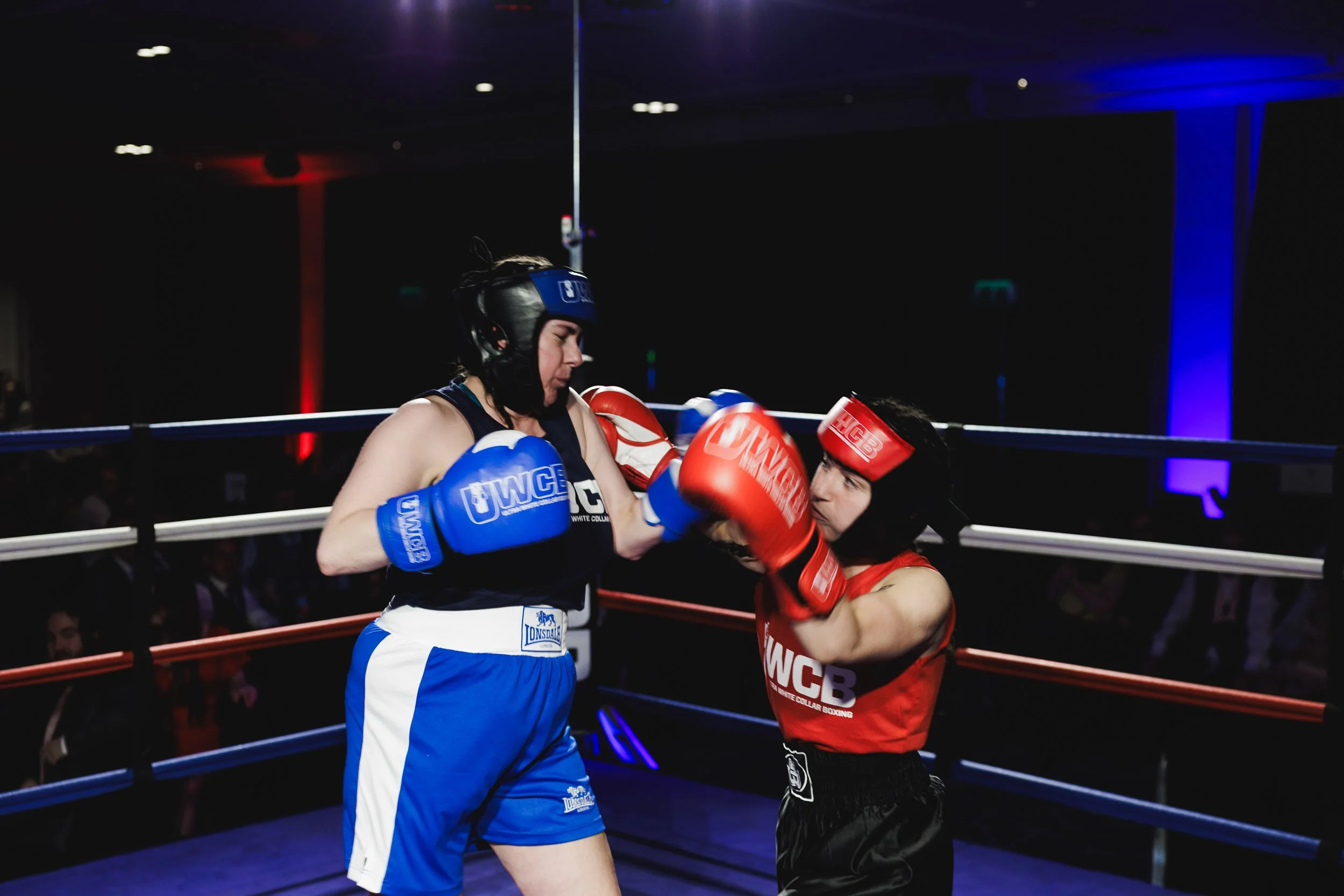 Two female boxers in a boxing ring, one in blue gear and one in red gear, engaged in a match with the boxer in red punching the boxer in blue.