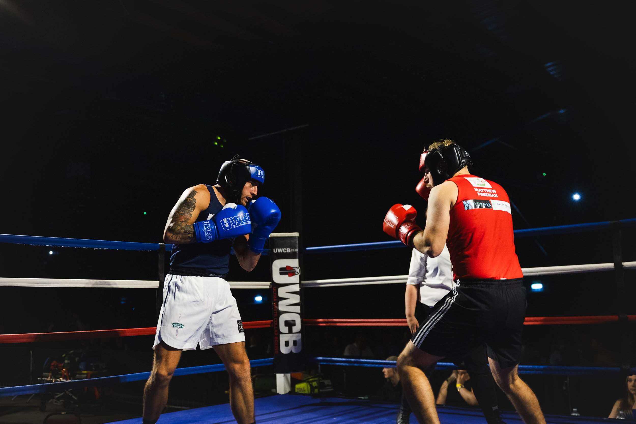 Boxing match between two male fighters in a ring, one in blue gloves and shorts, the other in red gloves and tank top, with a referee in the background.