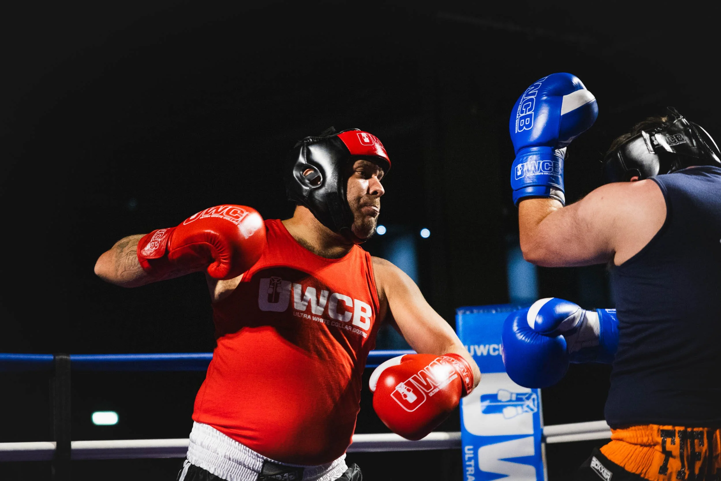 Two men in a boxing ring wearing protective helmets and boxing gloves, engaged in a fight, with a third person ready to strike.