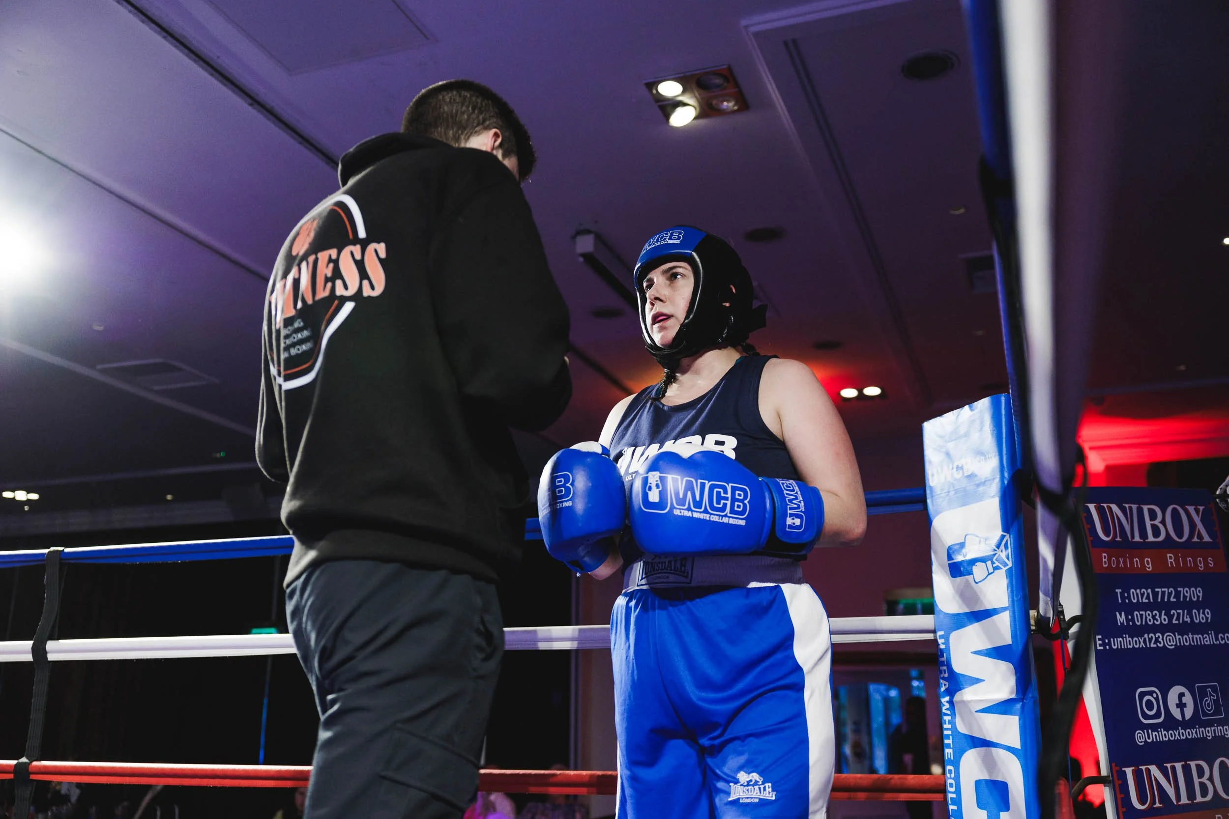 A female boxer with blue gloves and shorts standing in a ring, listening to her coach.