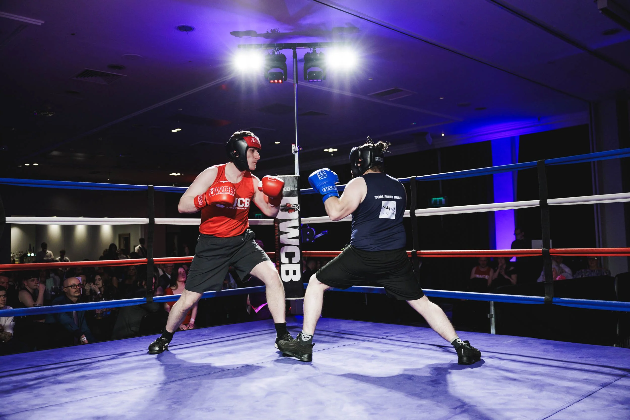 Two women boxing in a ring during a match, one in red gear and the other in blue gear, with spectators watching in the background.