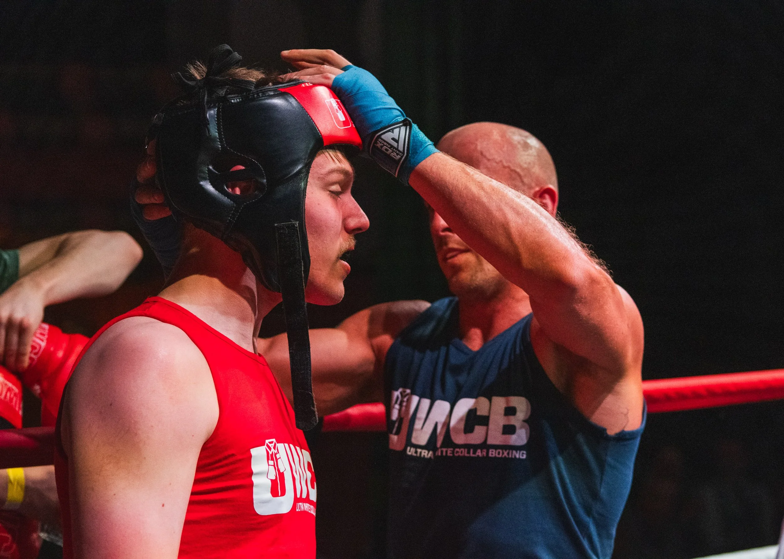 A boxing coach helps a young male boxer with his helmet in a boxing ring.